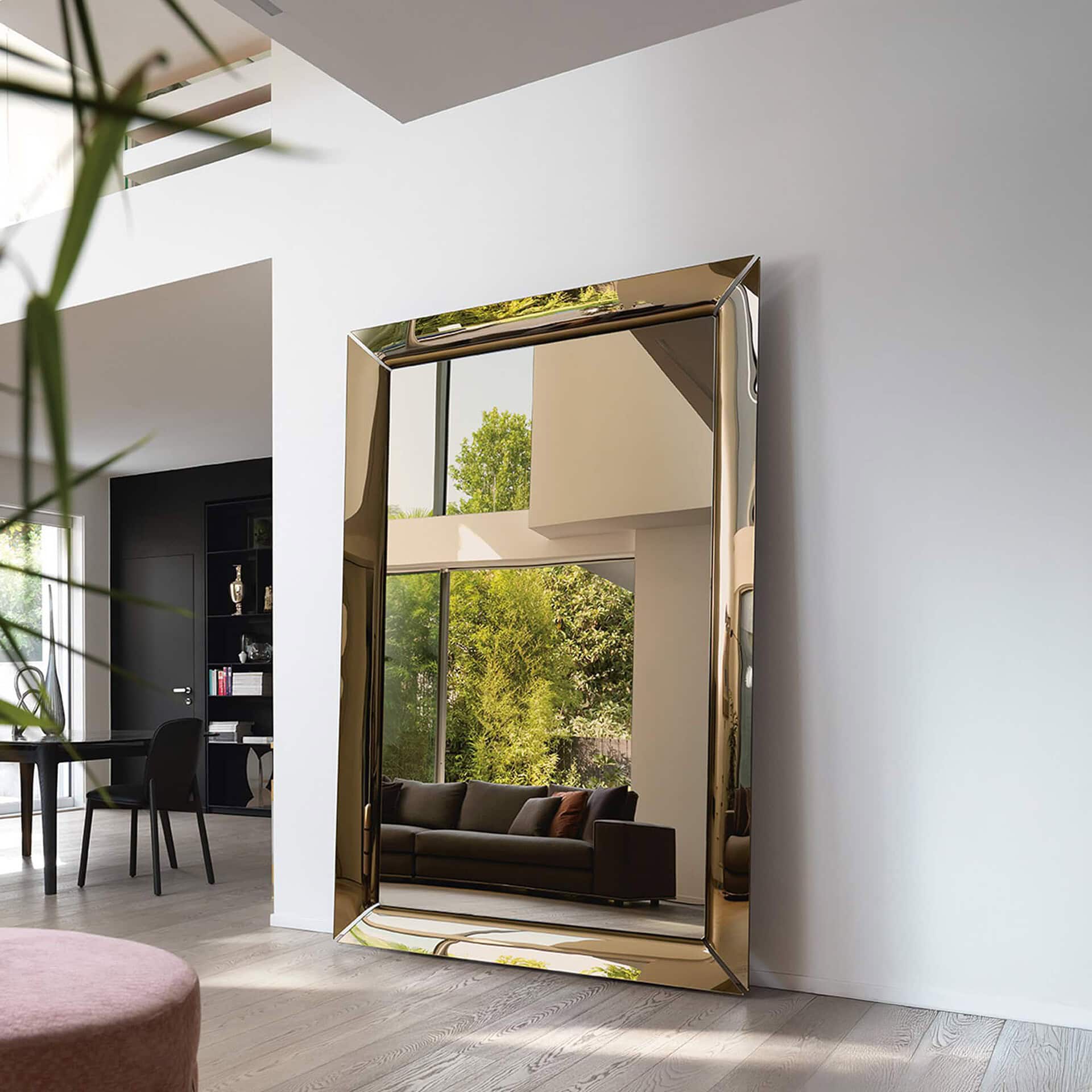 Caadre Mirror in champagne glass finish, leaning against a white wall in a modern living room with large windows reflecting greenery outside.