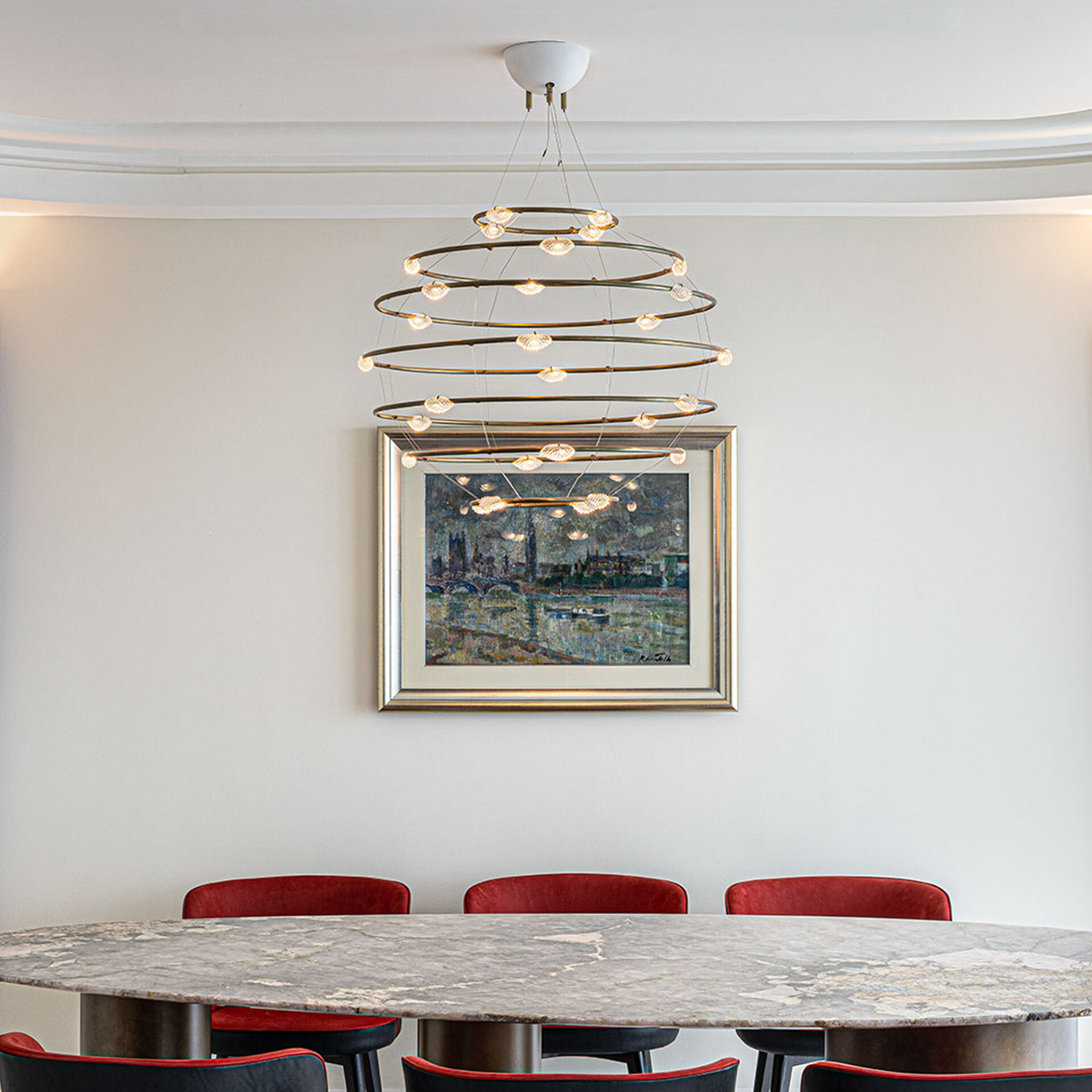 A modern dining room featuring a tiered ring chandelier, an impressionistic cityscape painting, a marble table, and red upholstered chairs.