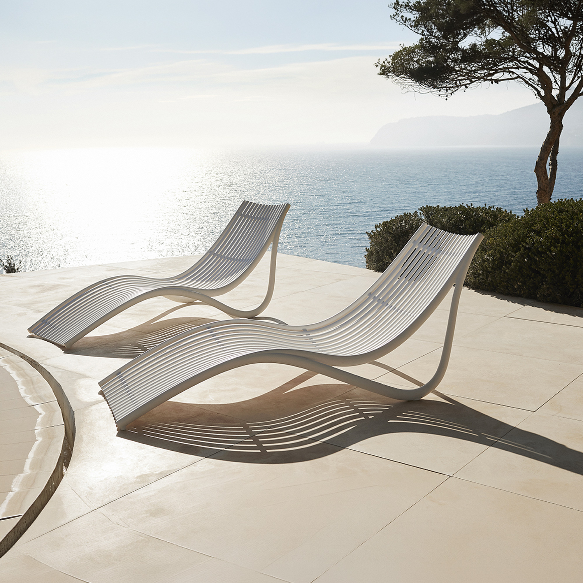 Two modern white slatted lounge chairs on a tiled patio overlooking a sparkling blue ocean under a bright sky.