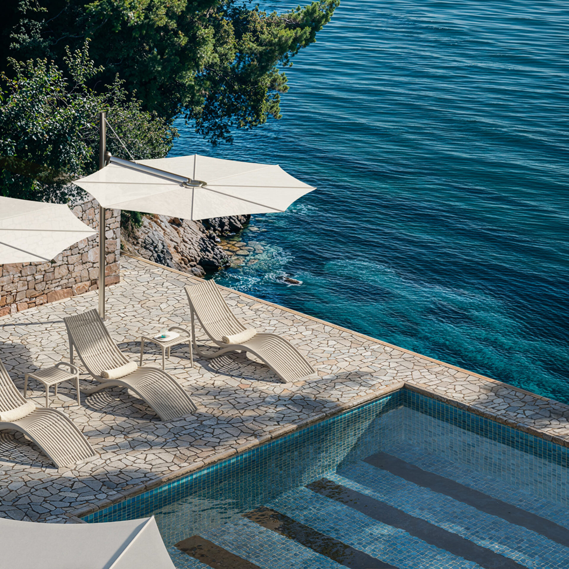 An aerial view of a luxurious outdoor patio with a swimming pool, modern lounge chairs under umbrellas, and a breathtaking view of the deep blue sea and rocky coastline.