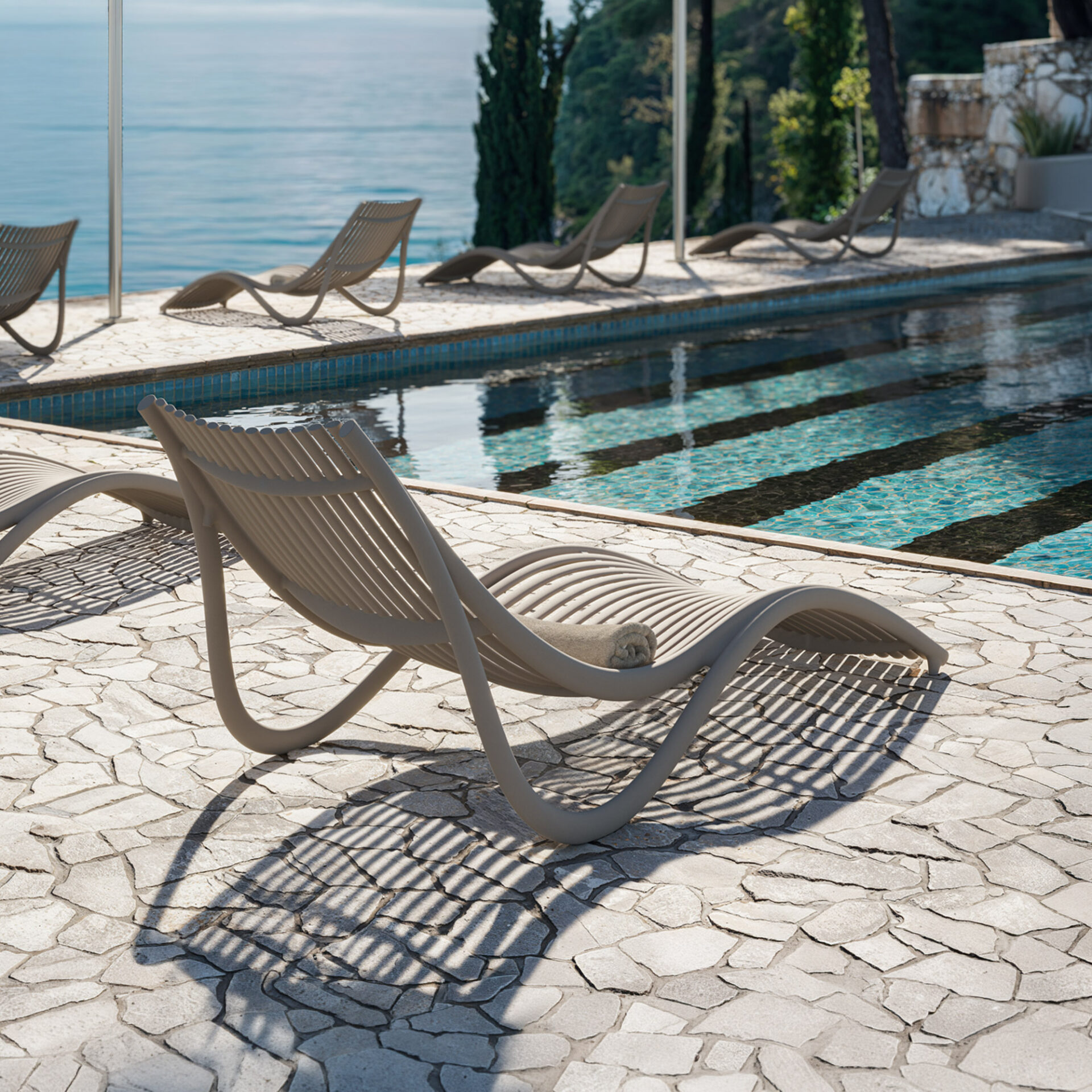 A modern, slatted lounge chair with a rolled towel on a flagstone patio next to a swimming pool, overlooking a vast blue ocean. Sunlight casts striped shadows.
