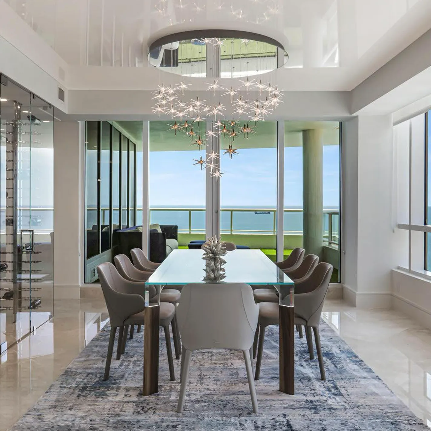 A modern dining room with a glass table, gray chairs, and a starburst chandelier, overlooking the ocean through floor-to-ceiling windows.