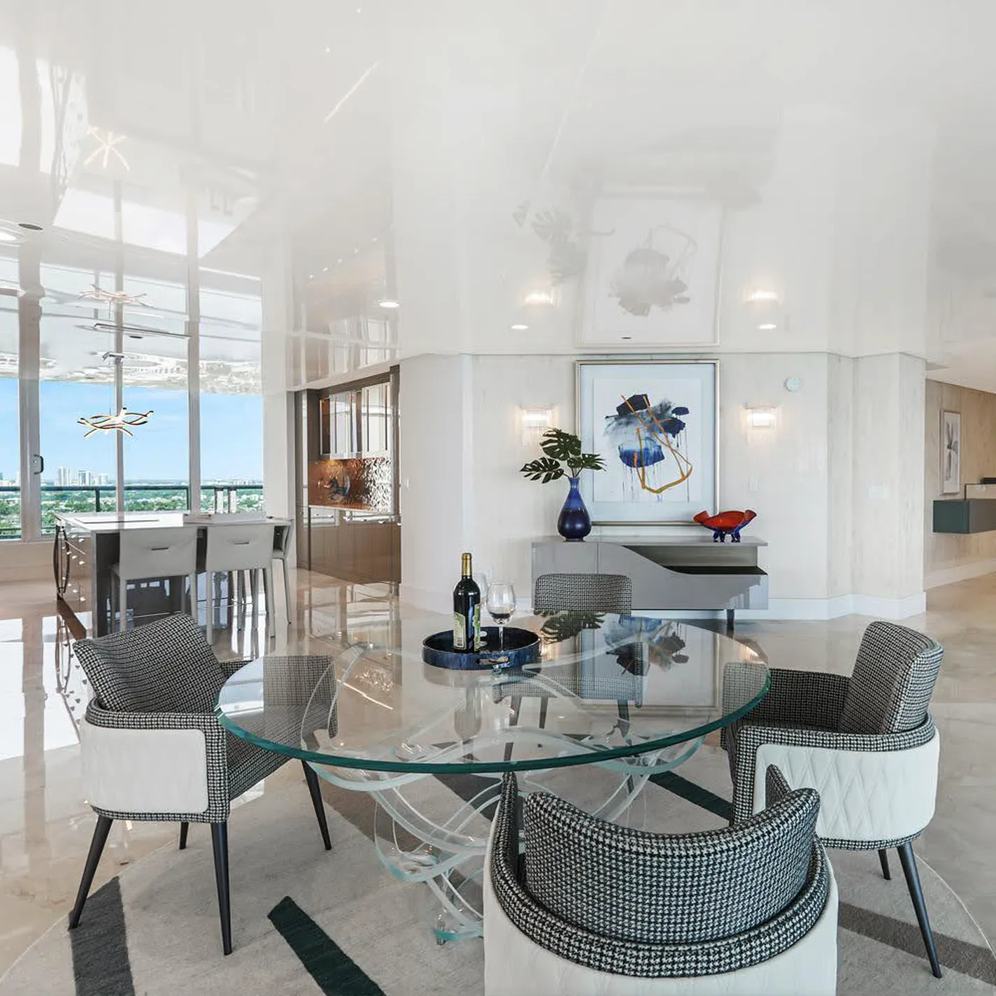 A luxurious modern dining room with a round glass table, four black and white houndstooth chairs, and large windows overlooking a city skyline. Abstract art and a console table adorn the background.