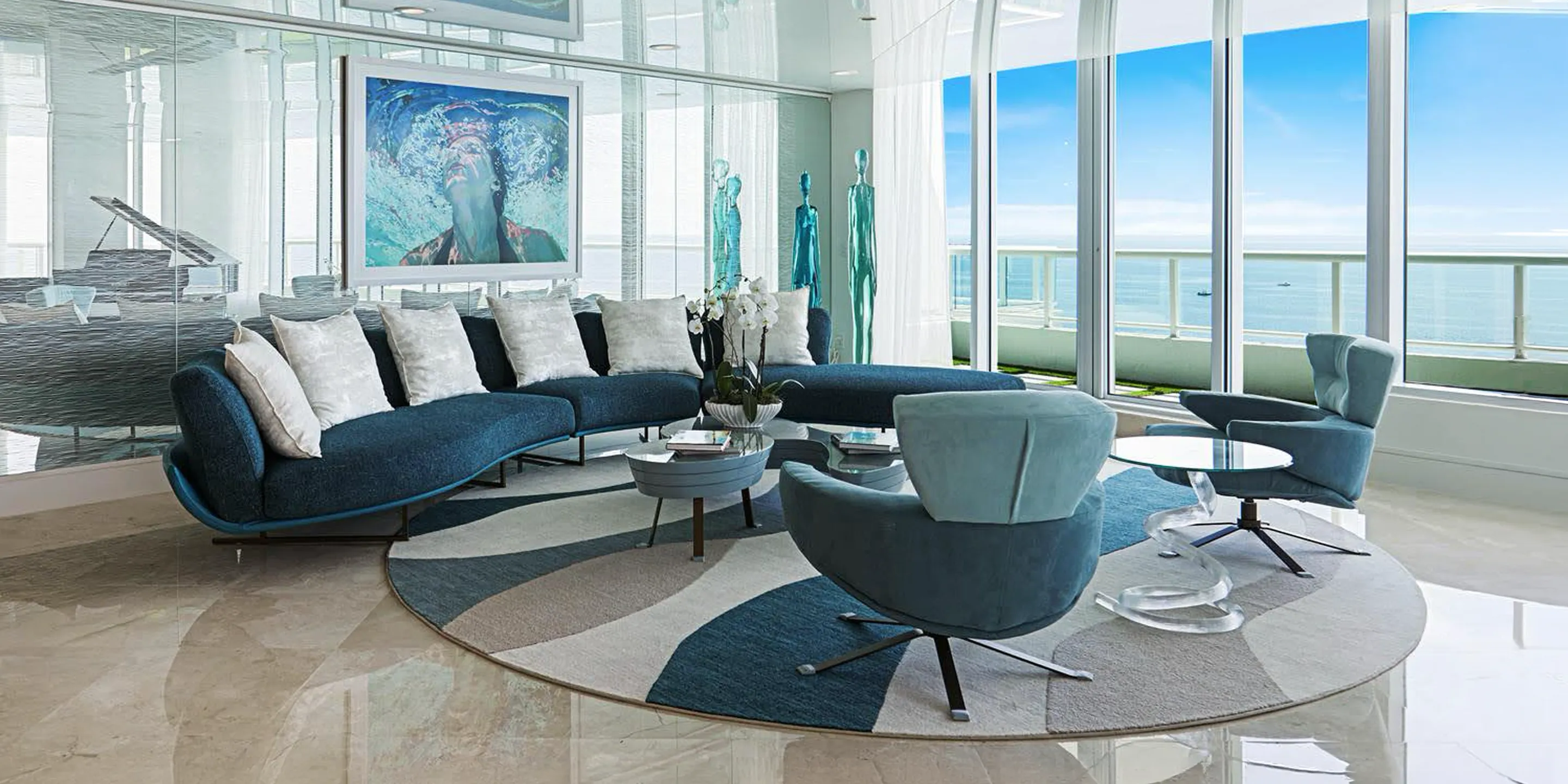 A modern living room with a curved blue sofa, armchairs, and a round rug, facing floor-to-ceiling windows with an ocean view.