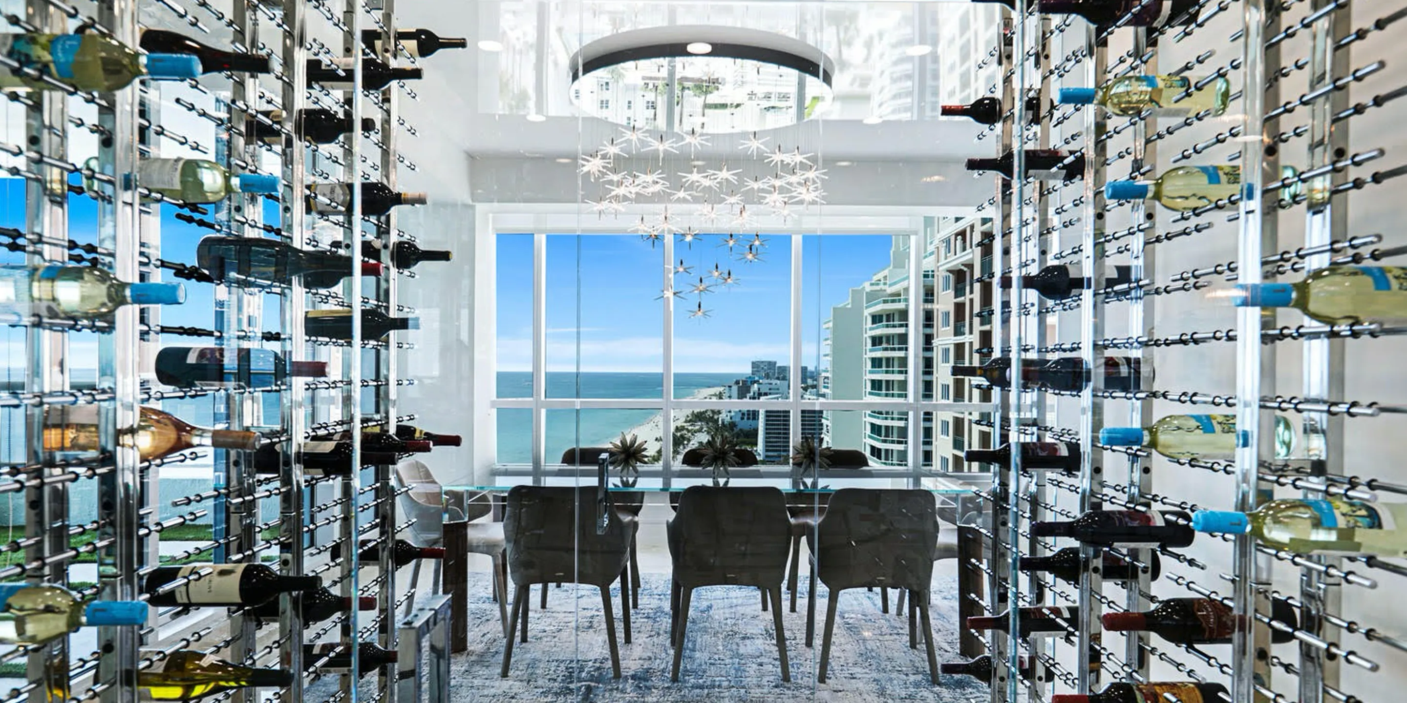 A modern dining room with floor-to-ceiling wine racks, a glass dining table, and a large window overlooking a beach, ocean, and city buildings.