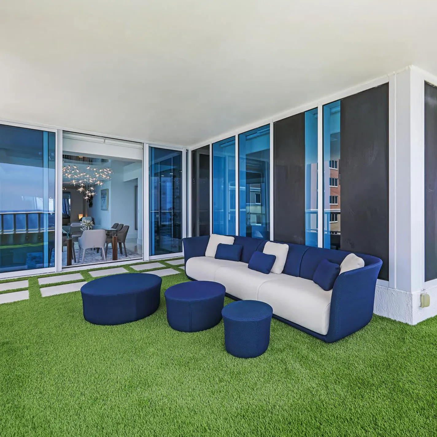 A modern balcony with green artificial turf, a blue and white sofa, and blue ottomans. Sliding glass doors lead to an indoor dining area with a starburst chandelier.