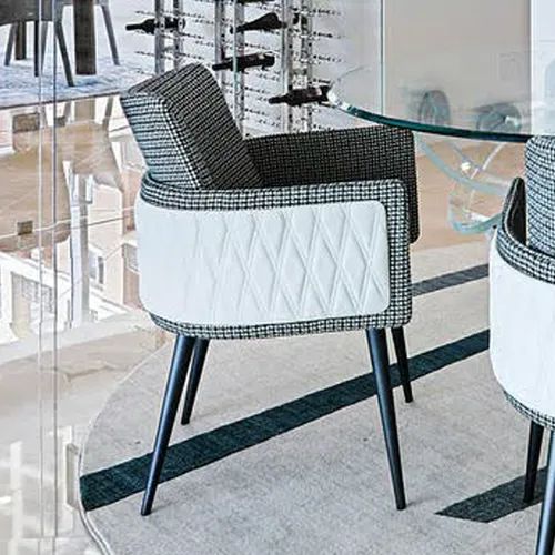 A modern dining chair with black and white houndstooth upholstery and white quilted leather panels, on a rug with a dark stripe, next to a glass table and a wine rack.