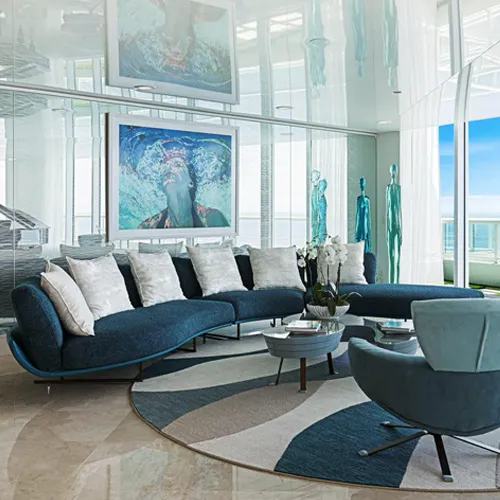 A modern living room with a curved blue sofa, a round striped rug, and two framed artworks of a person in water. Large windows offer a bright outdoor view.