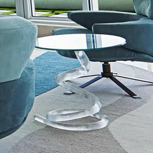 A modern living room with a round glass-top side table featuring a clear spiral acrylic base, positioned between two blue upholstered chairs on a patterned rug.