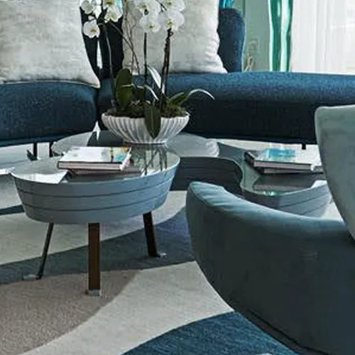 A modern living room with dark blue sofa, light blue armchair, two light blue coffee tables, an orchid plant, and a patterned rug.