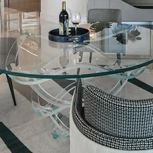 A round glass dining table with a sculptural clear base, set with a wine bottle and glass on a tray, next to a chair with a houndstooth pattern.