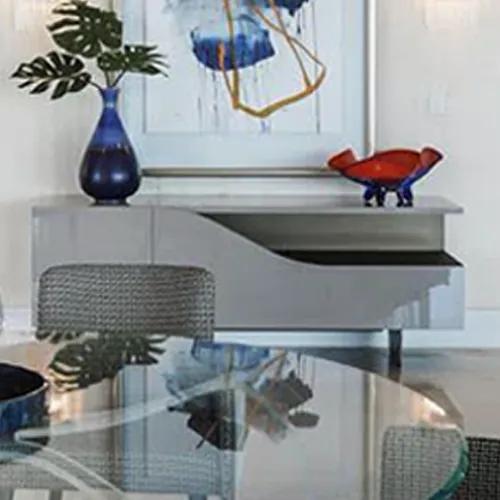 A modern grey console table with a blue vase of monstera leaves and a red and blue abstract sculpture, beneath an abstract painting.
