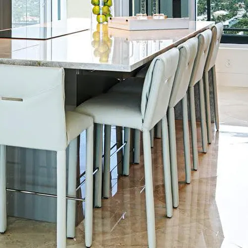 A modern kitchen island with a light-colored countertop and a row of cream-colored upholstered bar stools. A vase of green fruit sits on the island.