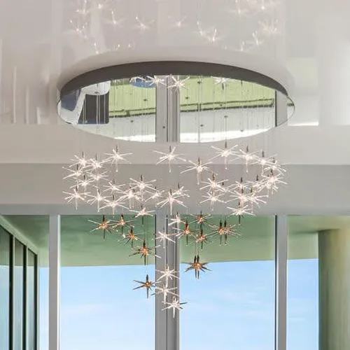 A modern chandelier with star-shaped lights arranged in a heart-like cascade, hanging from a mirrored ceiling fixture in front of large windows with a blue sky.