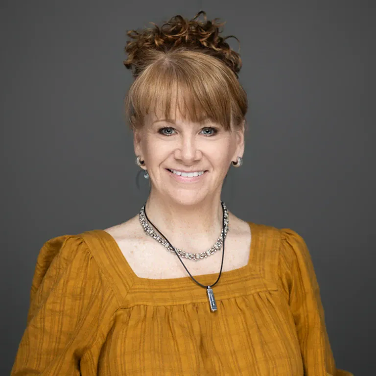 Portrait of a woman wearing a mustard blouse and jewelry against a gray background.