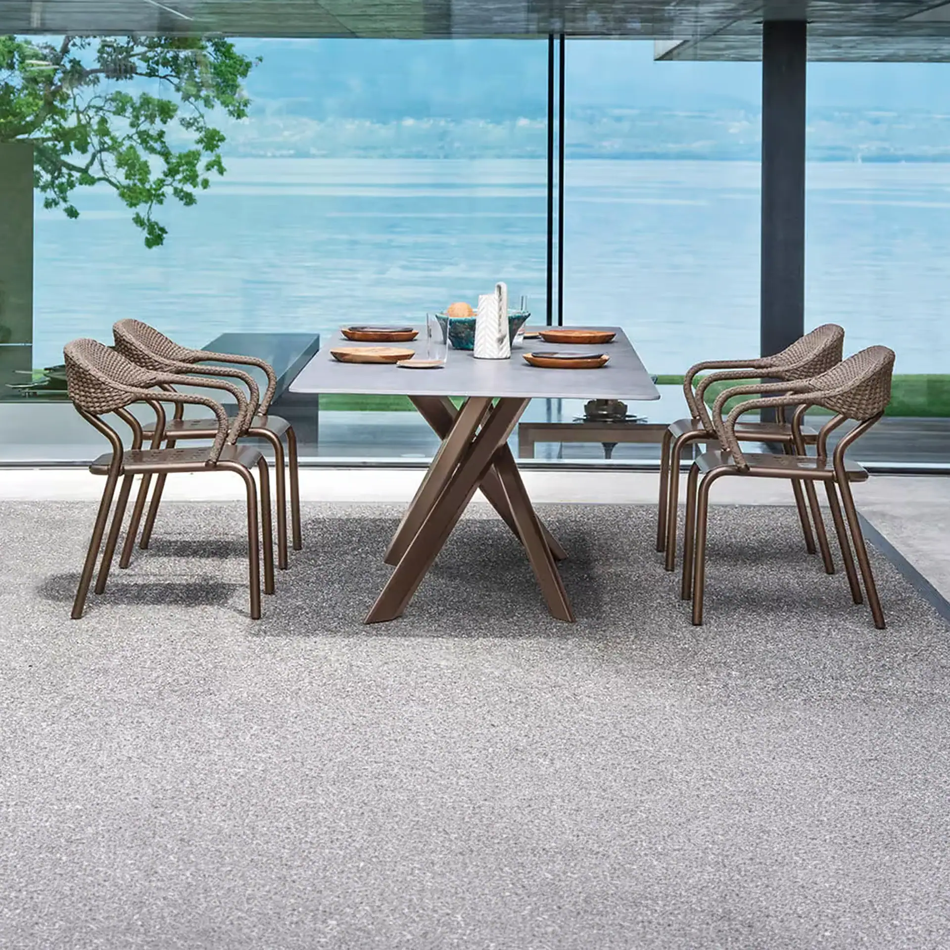 Modern outdoor dining table and four chairs with a gray rug, overlooking a large body of water through glass windows.