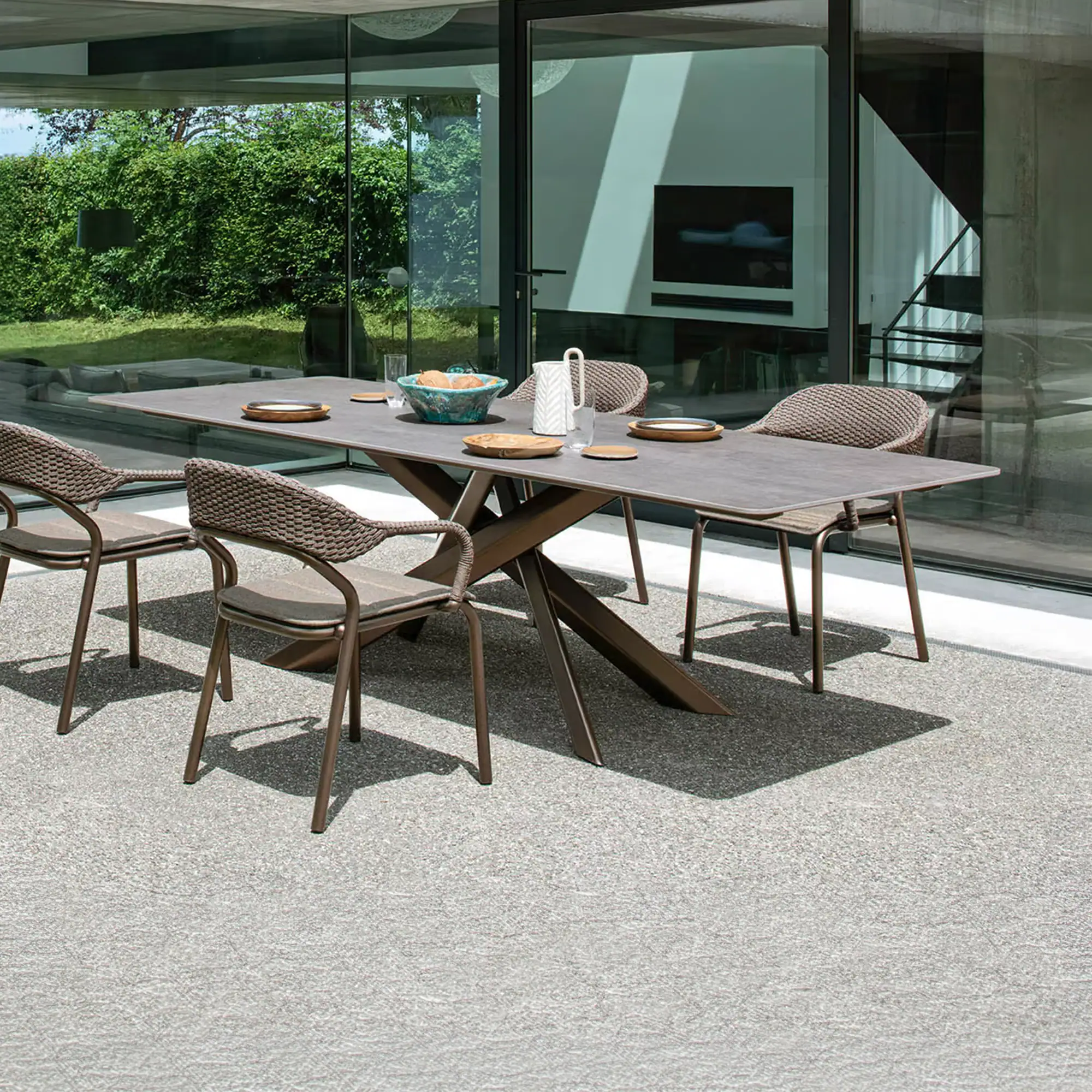 A modern outdoor dining set with a dark grey table and four brown woven chairs on a light grey patio, with glass doors reflecting a green garden and a modern interior.