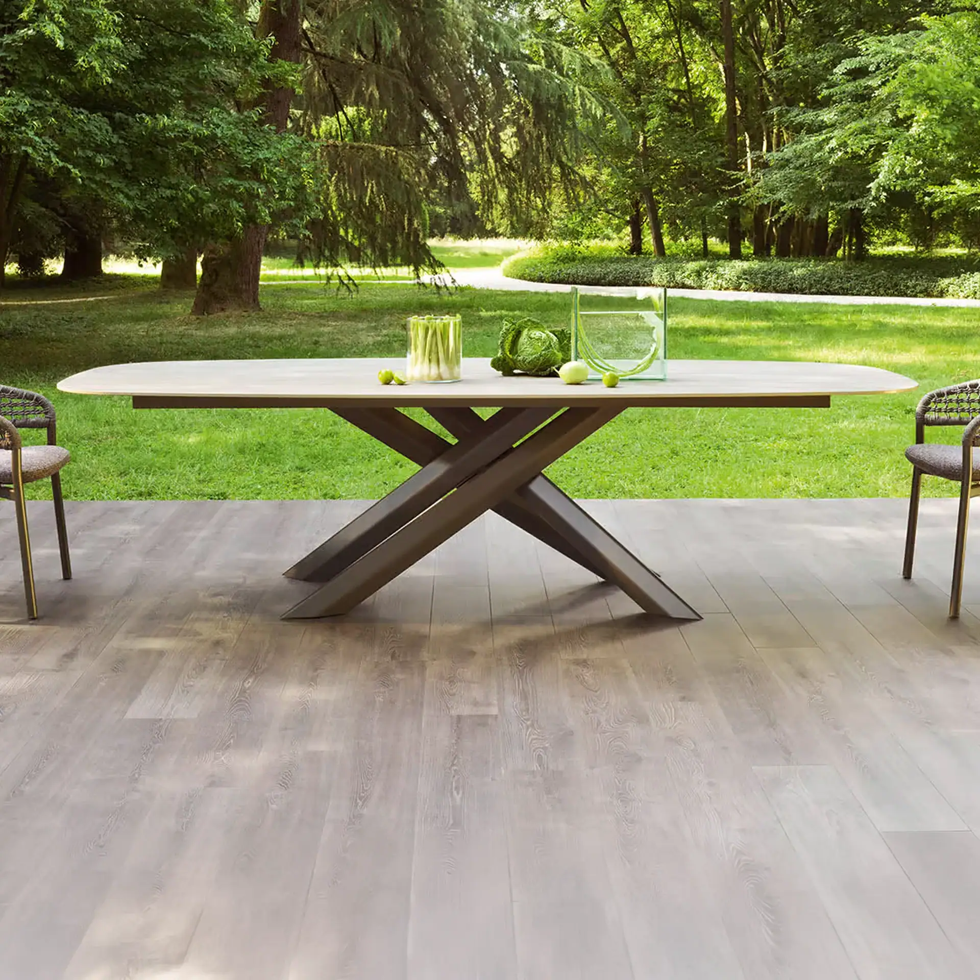 A modern oval outdoor dining table with a criss-cross base and two chairs on a wooden deck, surrounded by a vibrant green garden.
