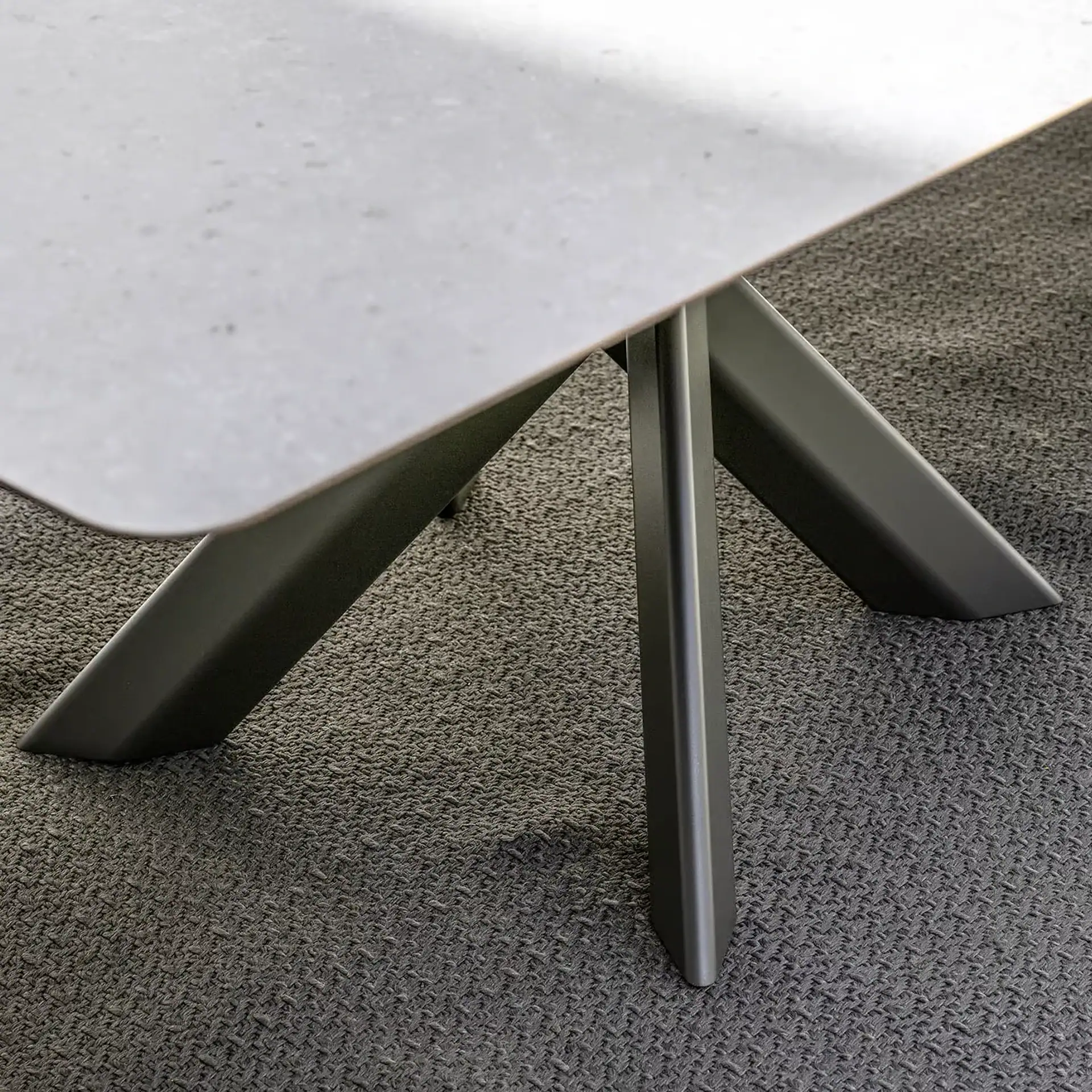 Close-up of a modern table's light gray surface and dark gray angled legs on a textured gray carpet.