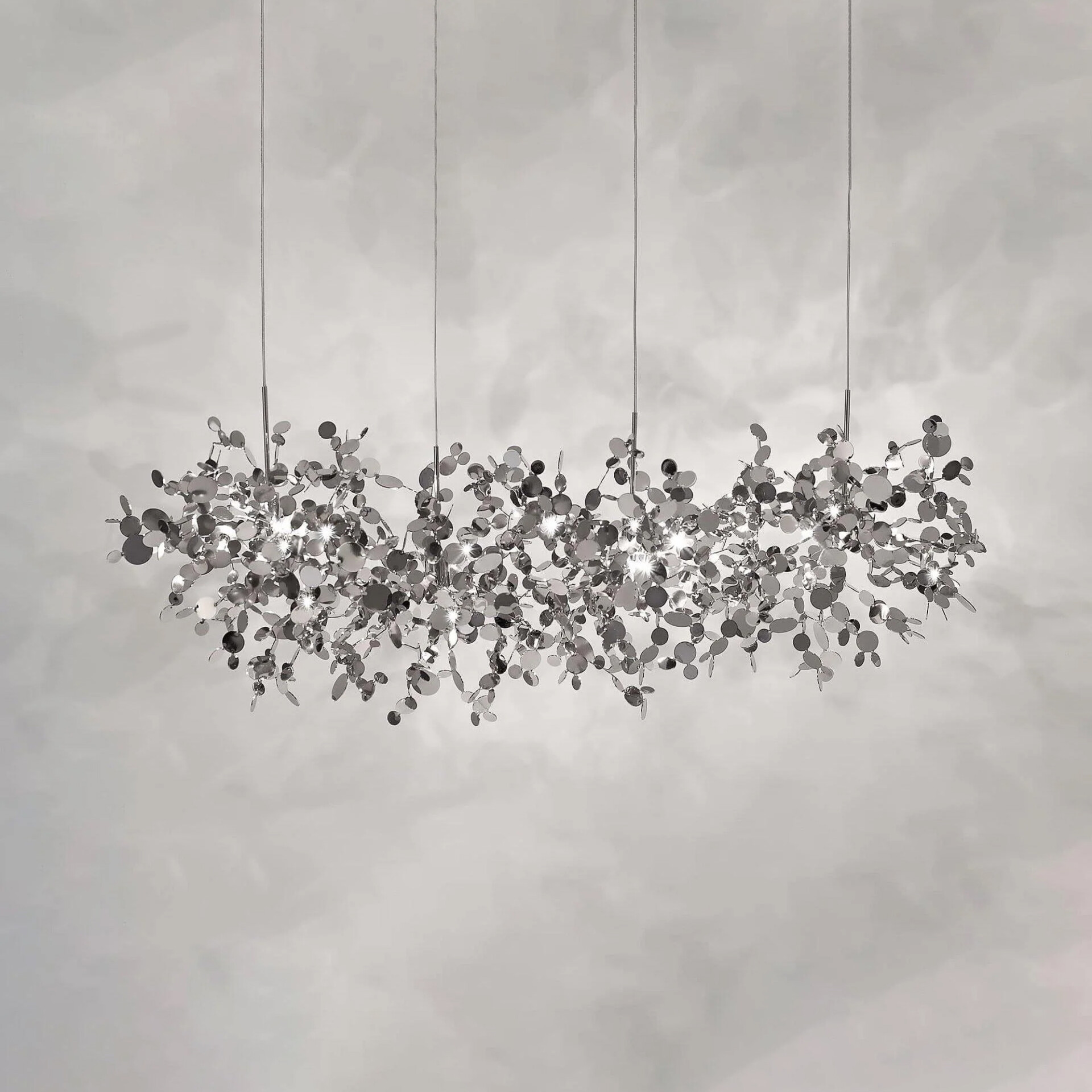 A modern, elongated chandelier made of numerous reflective silver discs, sparkling with light against a light gray background.