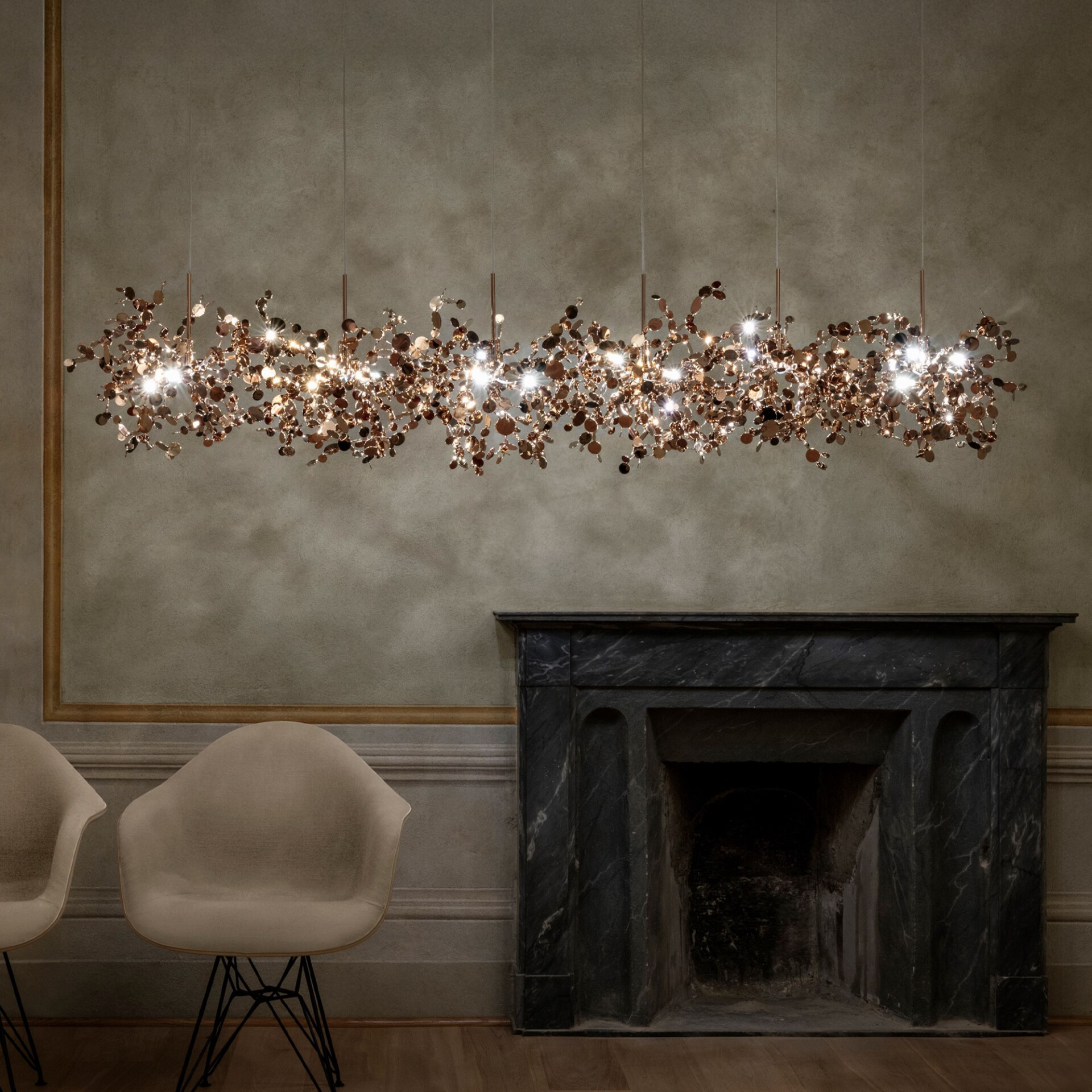 A long, linear chandelier made of numerous small, shimmering rose gold discs and bright lights hangs above a dark marble fireplace in an elegant room with muted walls and wooden floors.