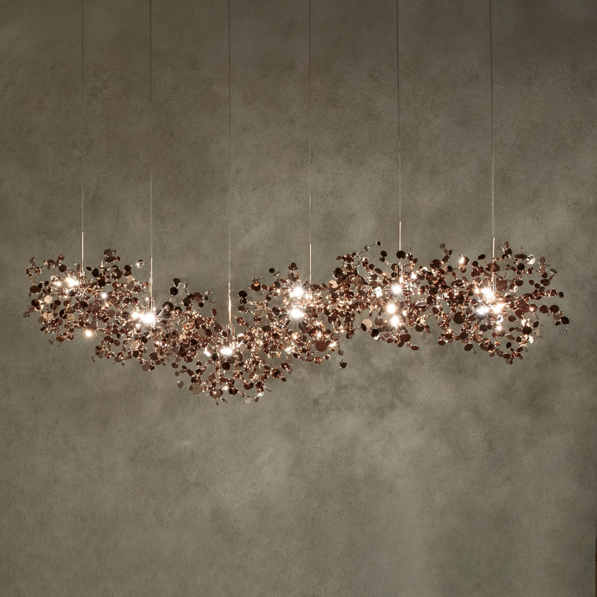 A long, organic-shaped chandelier made of many small, sparkling rose gold metallic discs, suspended against a dark, textured wall. Warm lights glow from within the fixture.