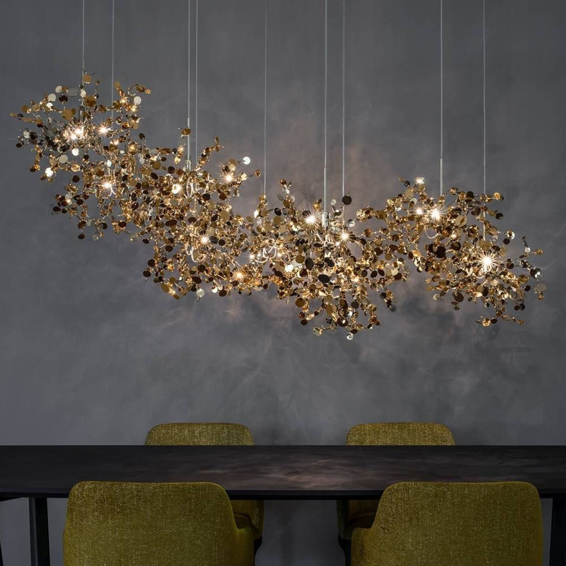 A large, intricate chandelier made of numerous gold and bronze metallic discs with integrated lights, suspended over a dark dining table with chartreuse chairs against a dark gray wall.