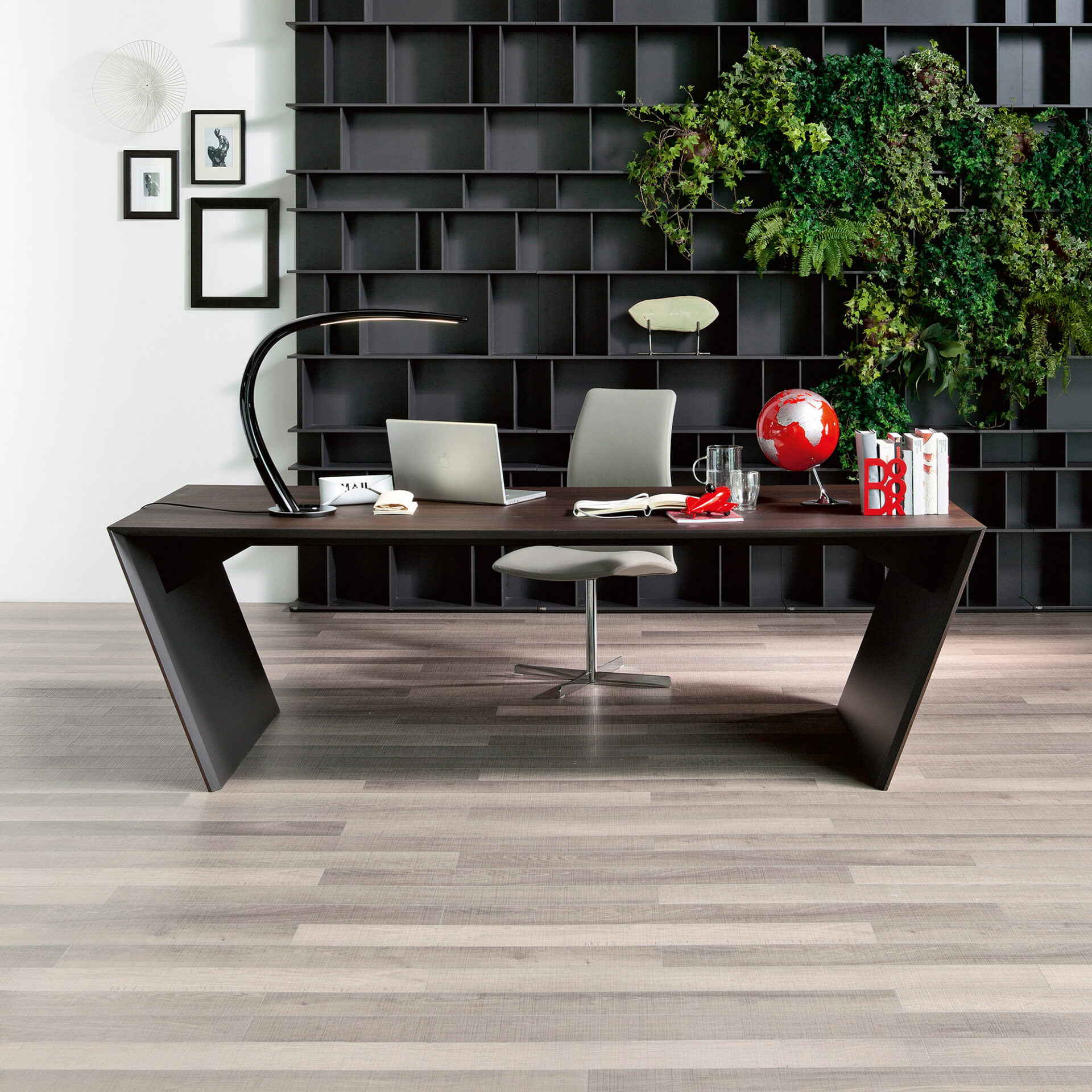 A modern home office with a dark brown desk, light grey office chair, and a large dark modular shelving unit adorned with a vibrant vertical garden of green plants. The desk holds a laptop, lamp, globe, and books.