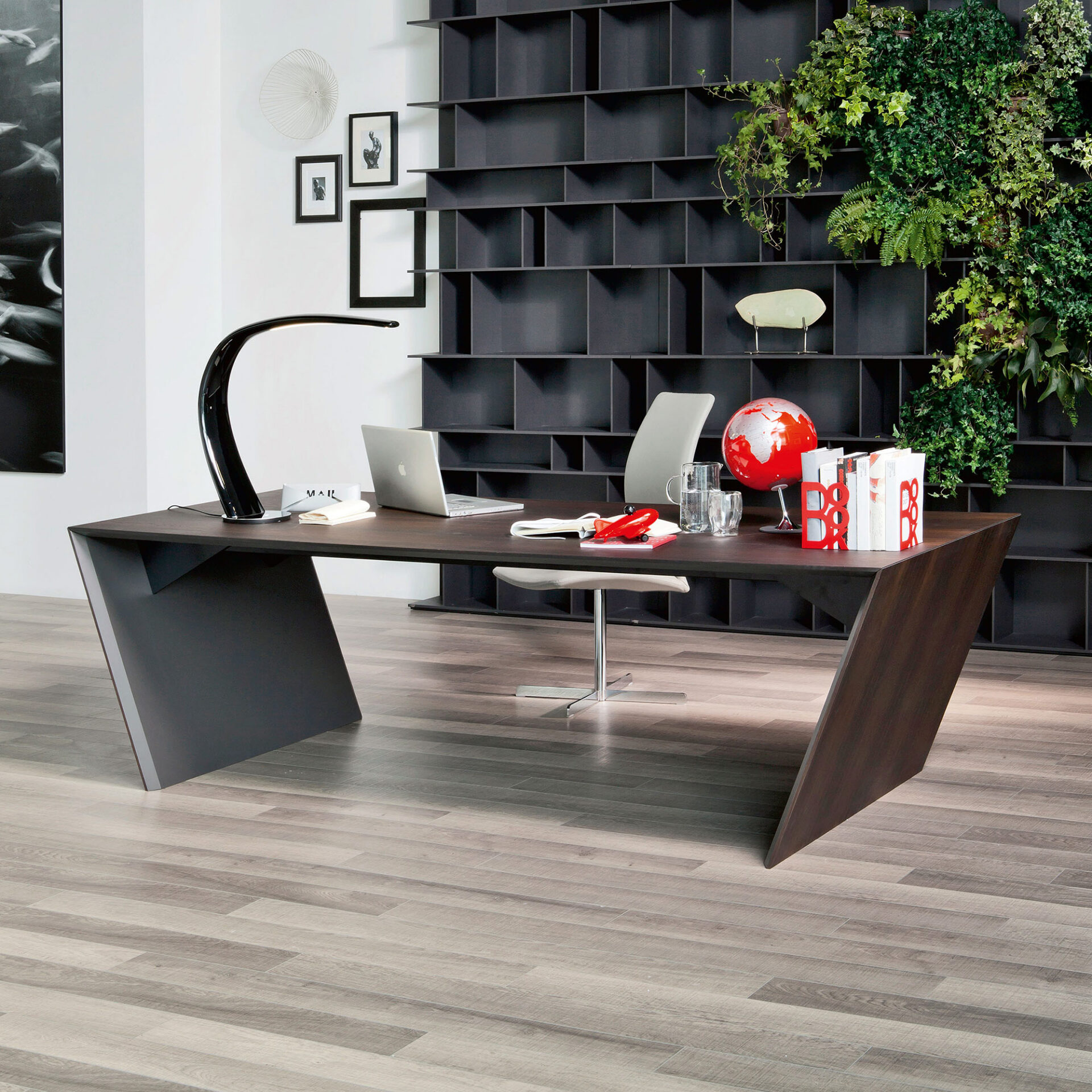 A modern home office with a dark, angular desk, a silver laptop, a black curved lamp, and a red globe. The background features a dark modular bookshelf with a lush green living wall on the right and framed art on a white wall to the left. The floor is light wood.