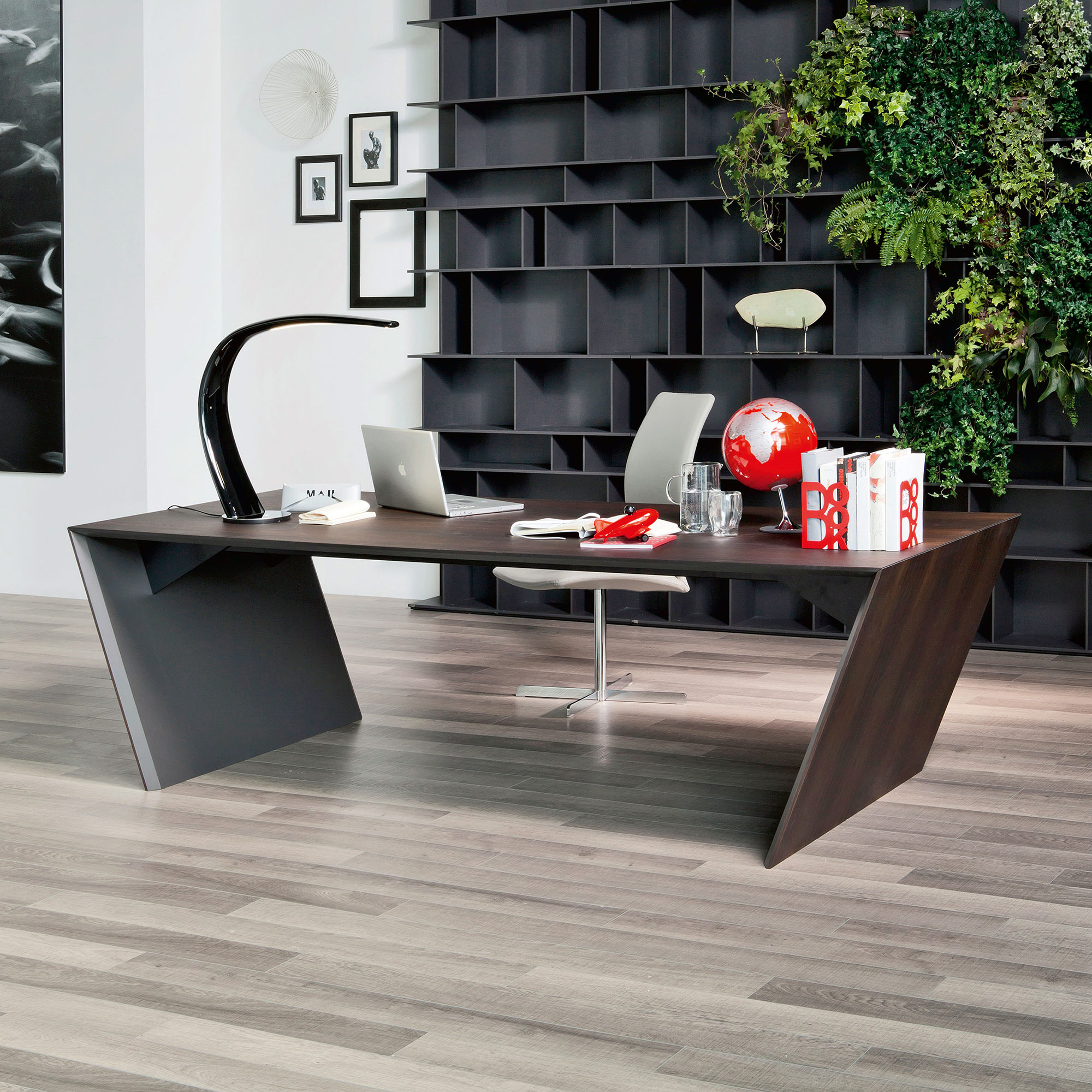 A modern home office with a dark, angular desk, a silver laptop, a black curved lamp, and a red globe. The background features a dark modular bookshelf with a lush green living wall on the right and framed art on a white wall to the left. The floor is light wood.