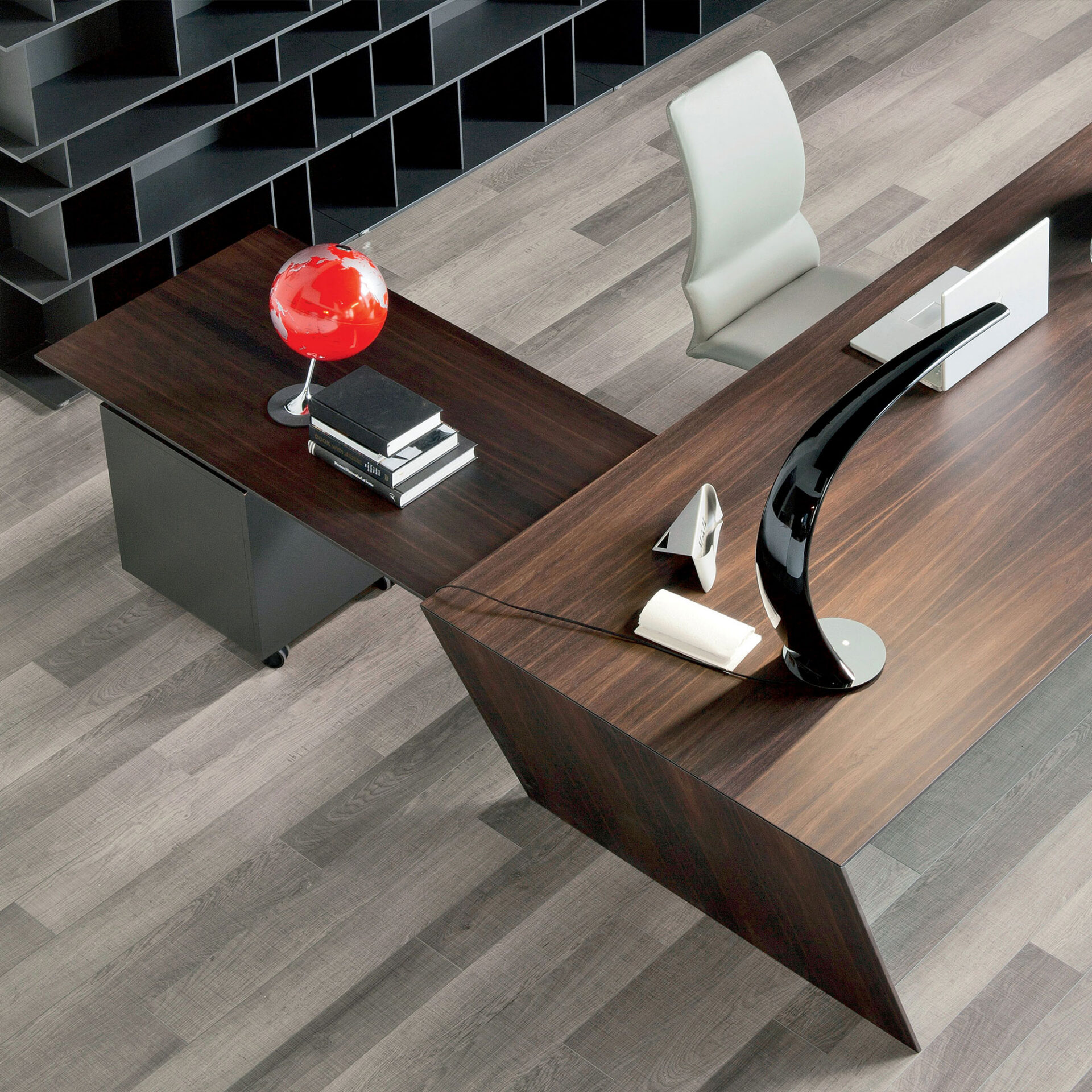 Overhead view of a modern L-shaped dark wood office desk with a light grey chair, red globe, black lamp, and dark modular shelving on grey wood flooring.