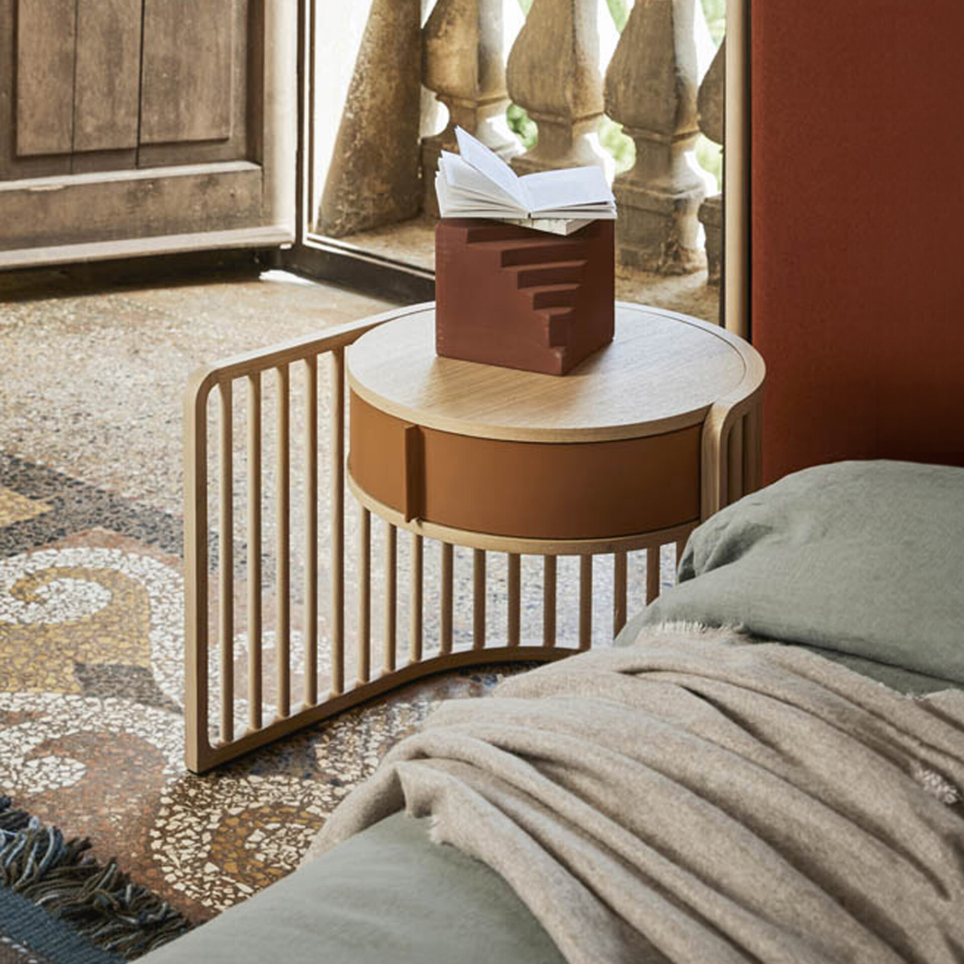 A modern round bedside table with a light wood top and terracotta drawer, featuring a slatted wooden base, sits beside a bed with a green duvet and beige throw. On the table is a terracotta step-shaped book stand holding an open book. A mosaic floor and an open window with a stone balustrade are in the background.