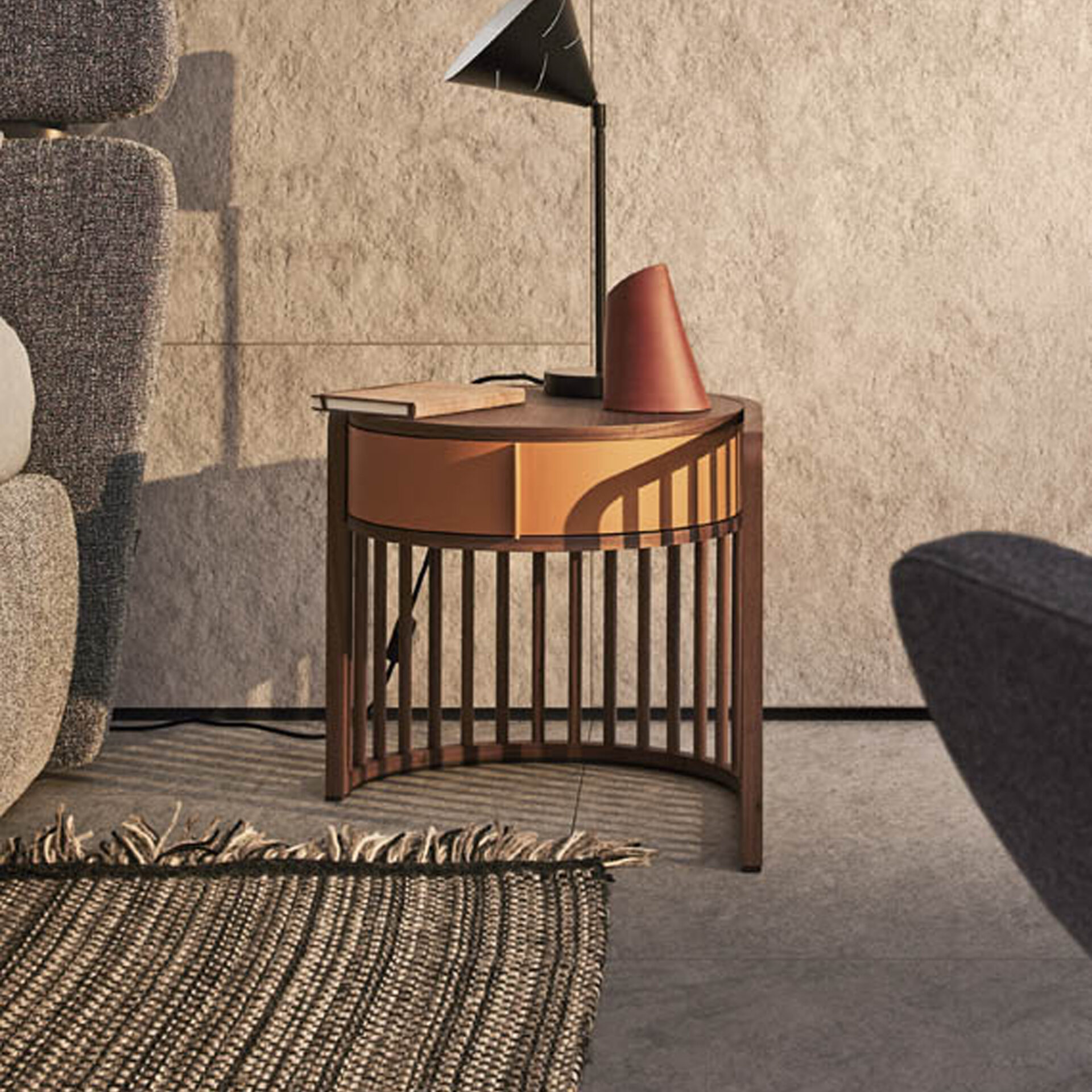 A modern, semi-circular wooden side table with an orange drawer and slatted base, topped with a black conical lamp and a book, set against a textured beige wall with strong shadows. A woven rug is in the foreground, and parts of grey upholstered chairs are visible on either side.