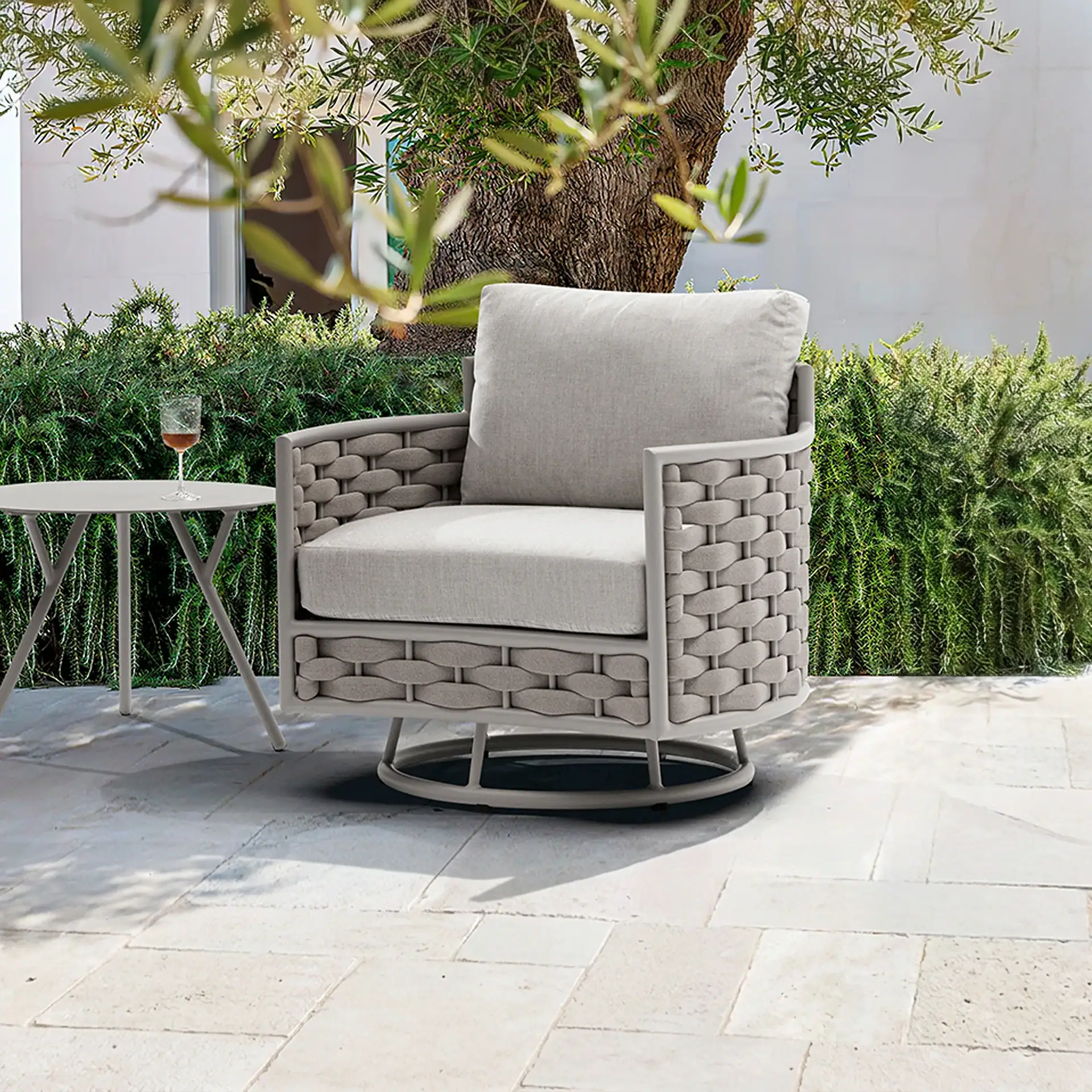 A gray woven outdoor armchair with light cushions and a small side table with a wine glass on a stone patio, surrounded by green hedges and a tree.