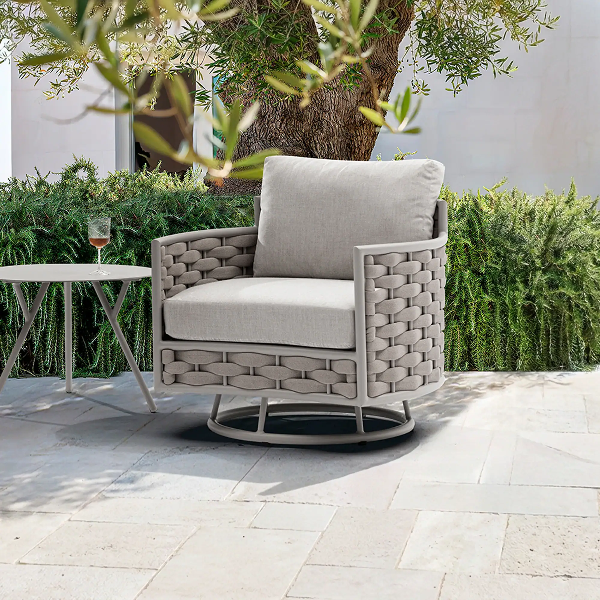 A gray woven outdoor armchair with light cushions and a small side table with a wine glass on a stone patio, surrounded by green hedges and a tree.