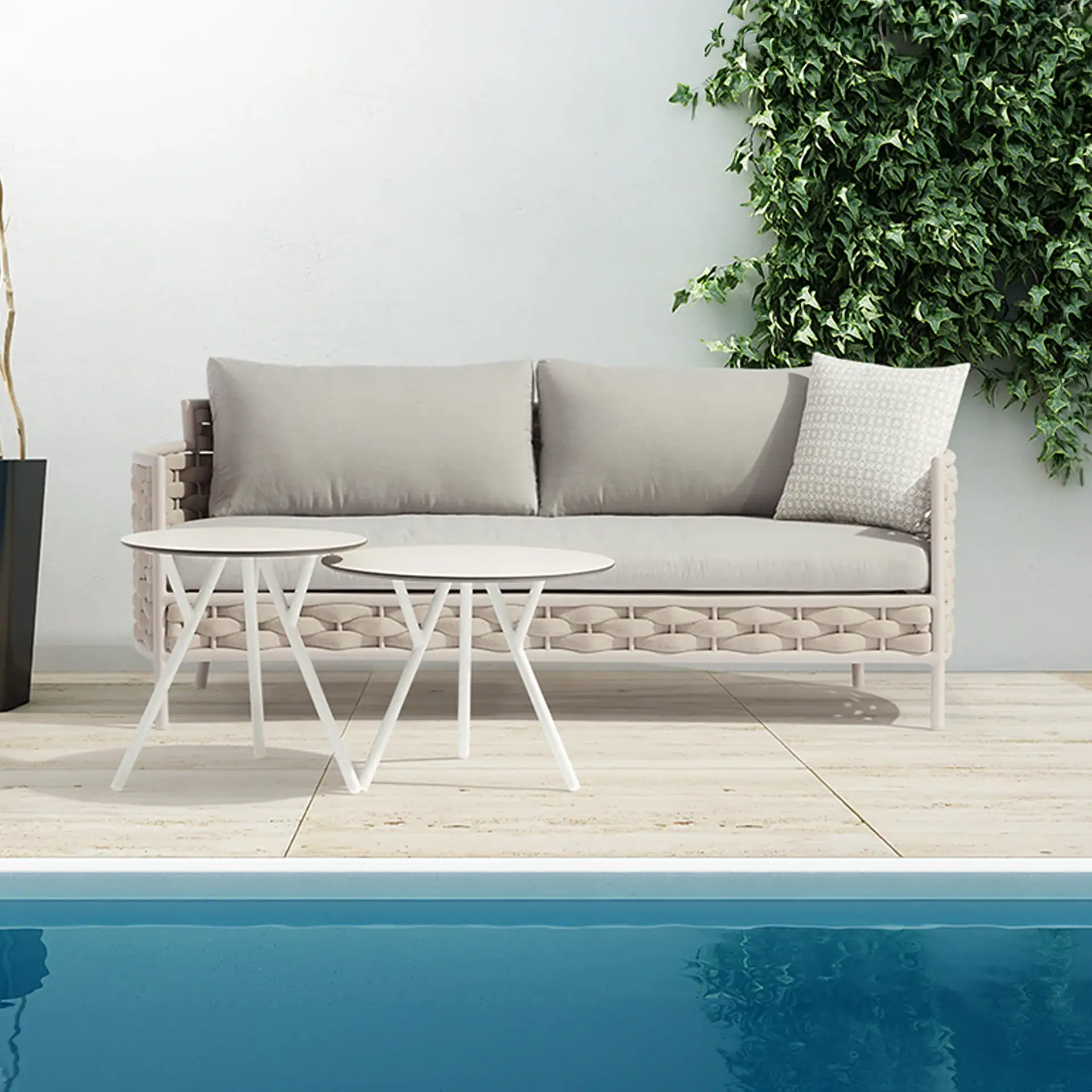 An outdoor patio with a light grey sofa, two white side tables, and a swimming pool in the foreground. Green ivy covers a white wall behind the sofa.
