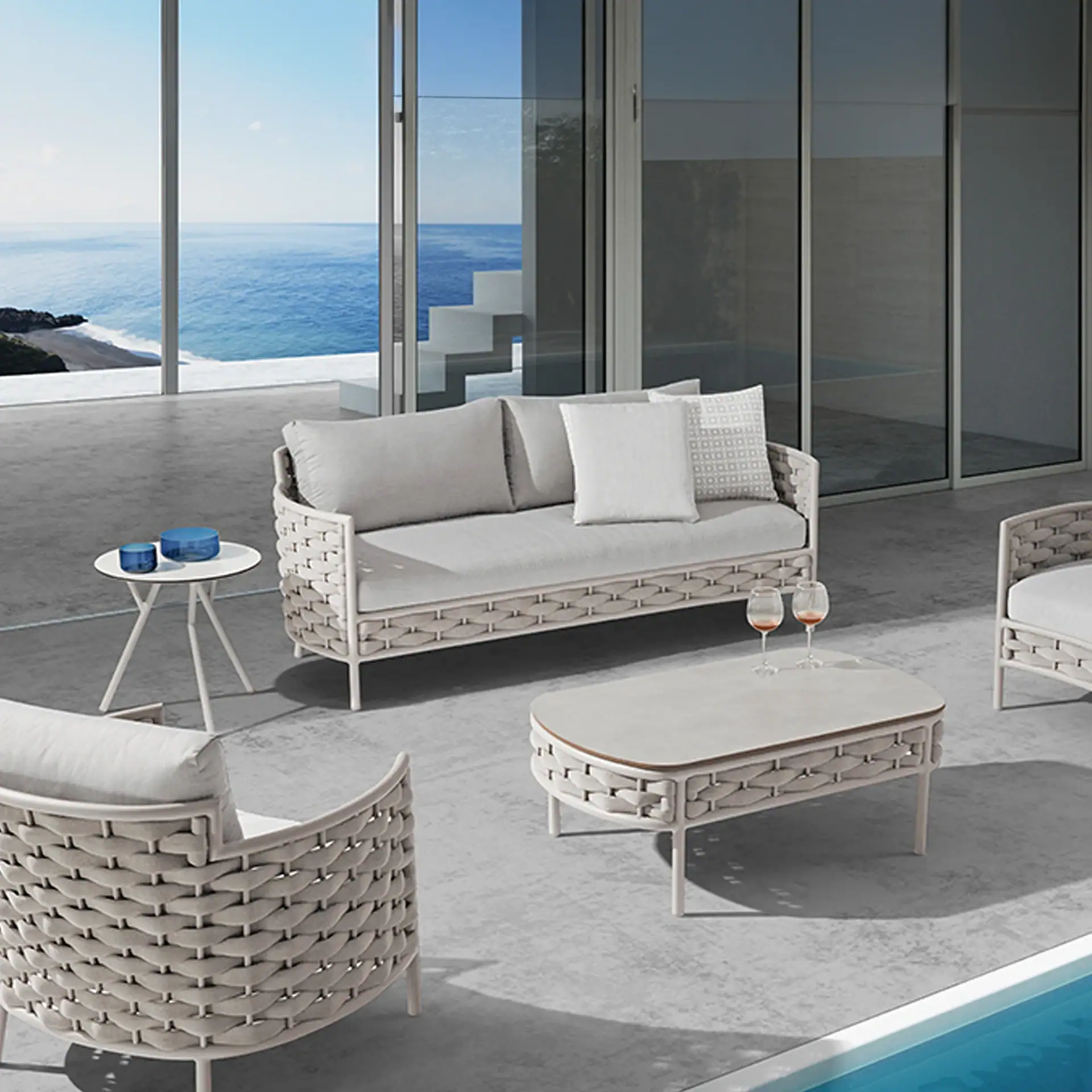 A luxurious outdoor patio with modern woven furniture, including a sofa, armchairs, coffee table, and side table, overlooking a blue ocean and a swimming pool.