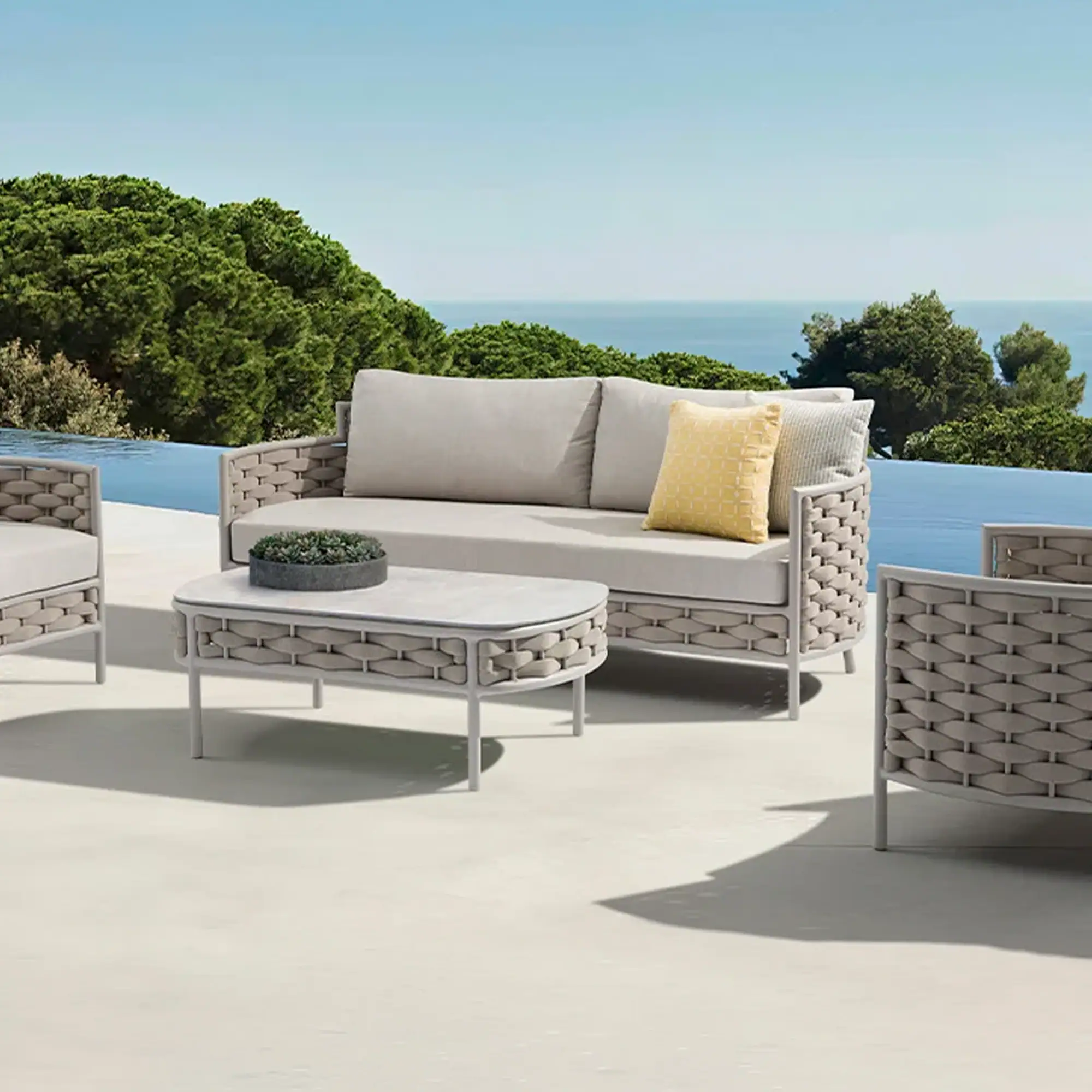 A modern light gray outdoor sofa, two armchairs, and an oval coffee table with woven details on a patio overlooking an infinity pool, green trees, and the ocean under a clear blue sky.
