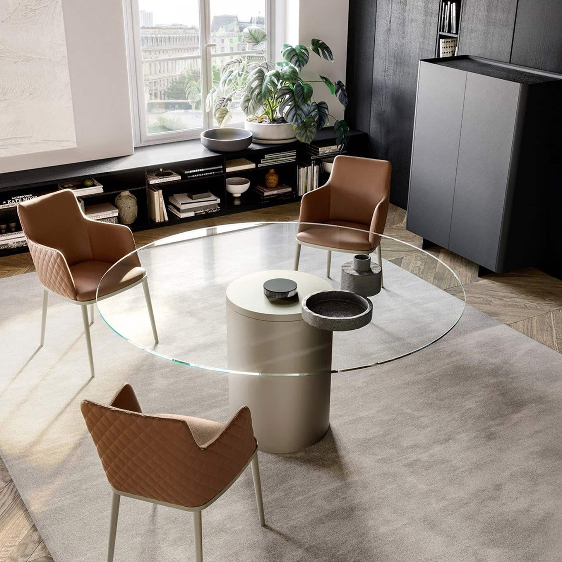 A modern dining room with a round glass table, four brown quilted chairs, a large window with a city view, and dark shelving with decor.