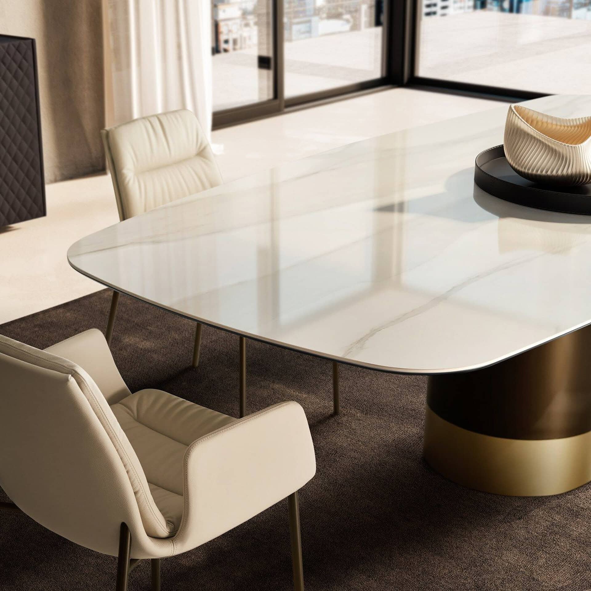 A modern dining room featuring a white marble table with a two-tone pedestal base, surrounded by cream leather chairs. Large windows in the background offer a blurred city view.