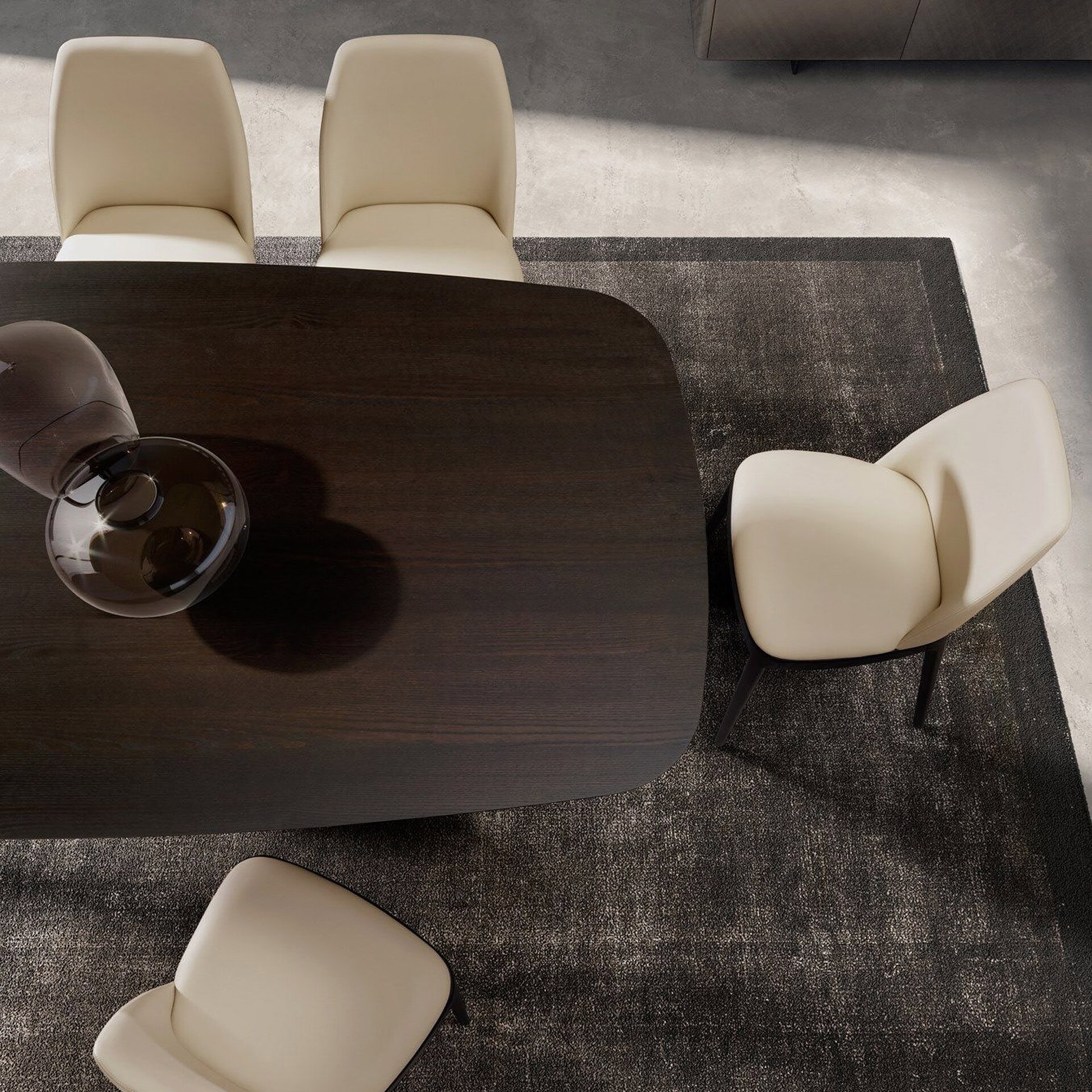 Overhead view of a modern dining room featuring a dark wood table, cream upholstered chairs, and a dark textured rug on a light gray concrete floor.