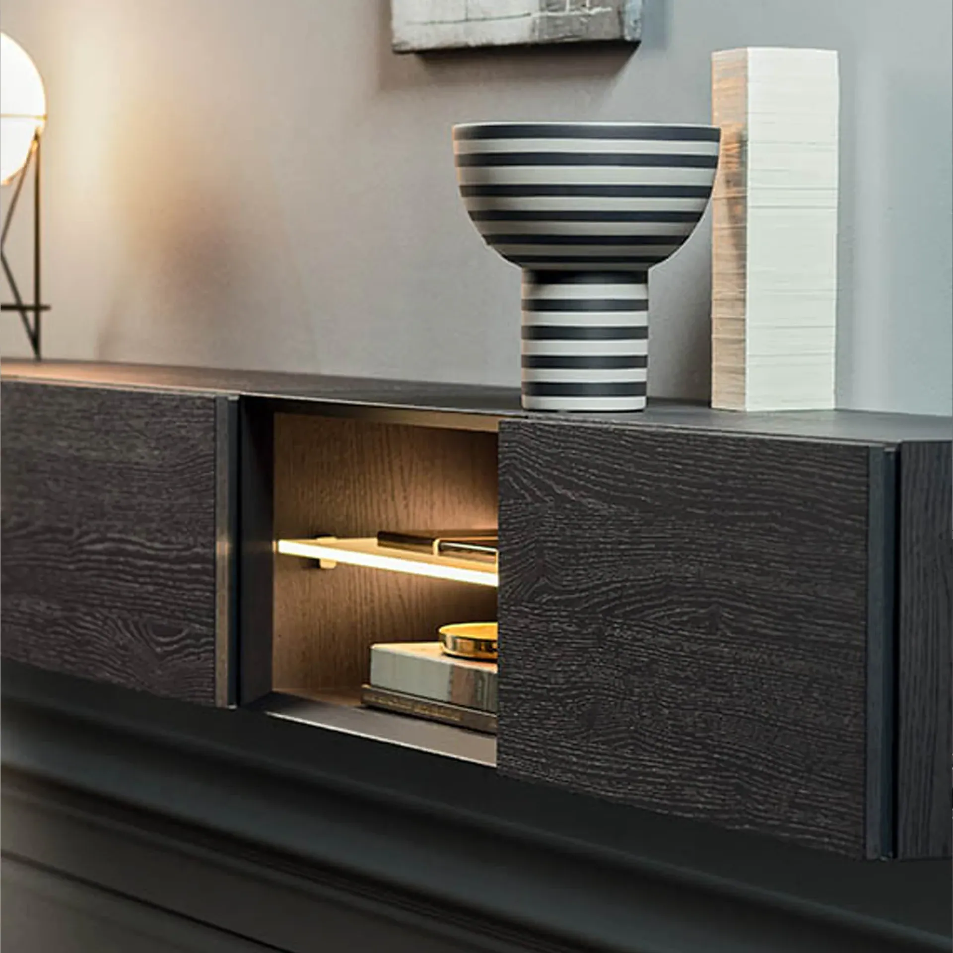A dark wood-grain wall-mounted cabinet with a partially open, illuminated section. A striped vase and white decorative blocks are on top.