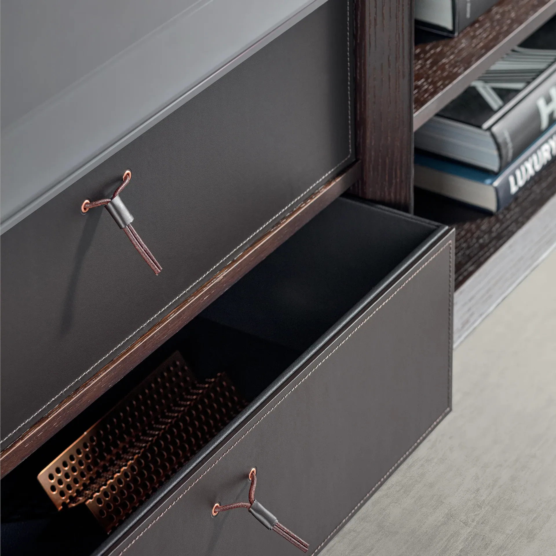 Two dark brown leather drawers with light stitching and leather cord handles; one drawer is open, revealing a copper perforated tray. Books are visible on a shelf to the right.