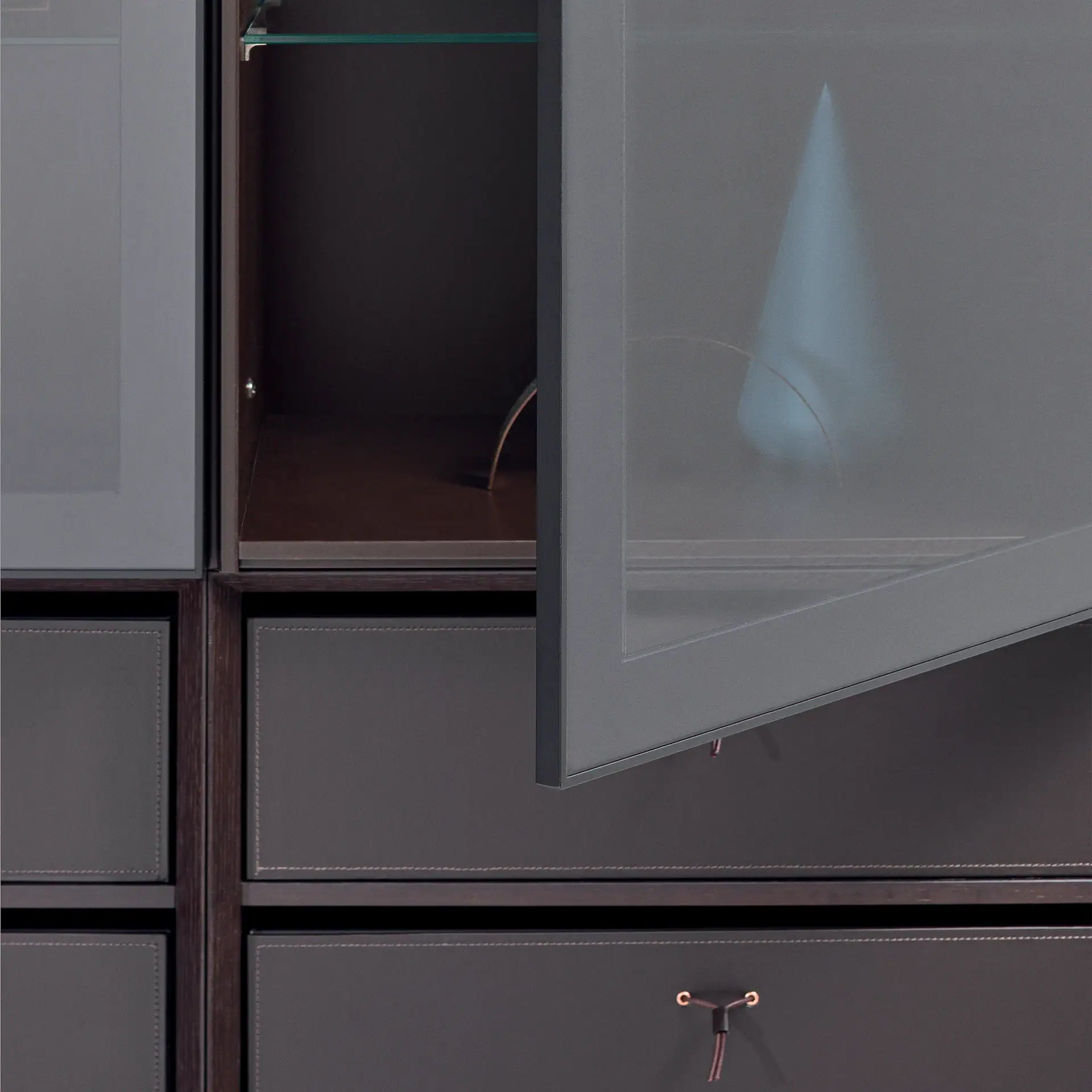 Close-up of a modern storage unit with dark gray translucent glass cabinets and dark gray leather-look drawers with light stitching and copper cord handles.