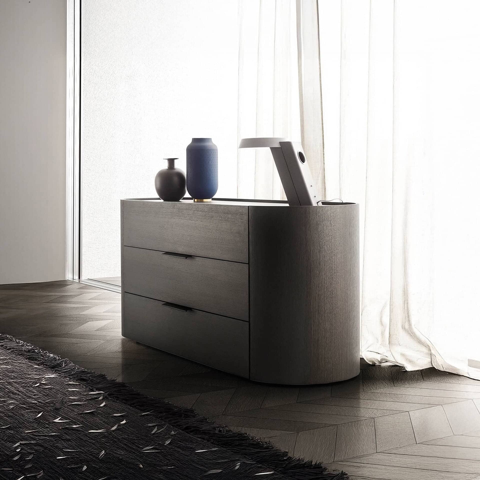 A dark wood dresser with rounded corners and three drawers, topped with two decorative vases and a pop-up light/screen. It sits on a dark herringbone wood floor next to sheer white curtains.
