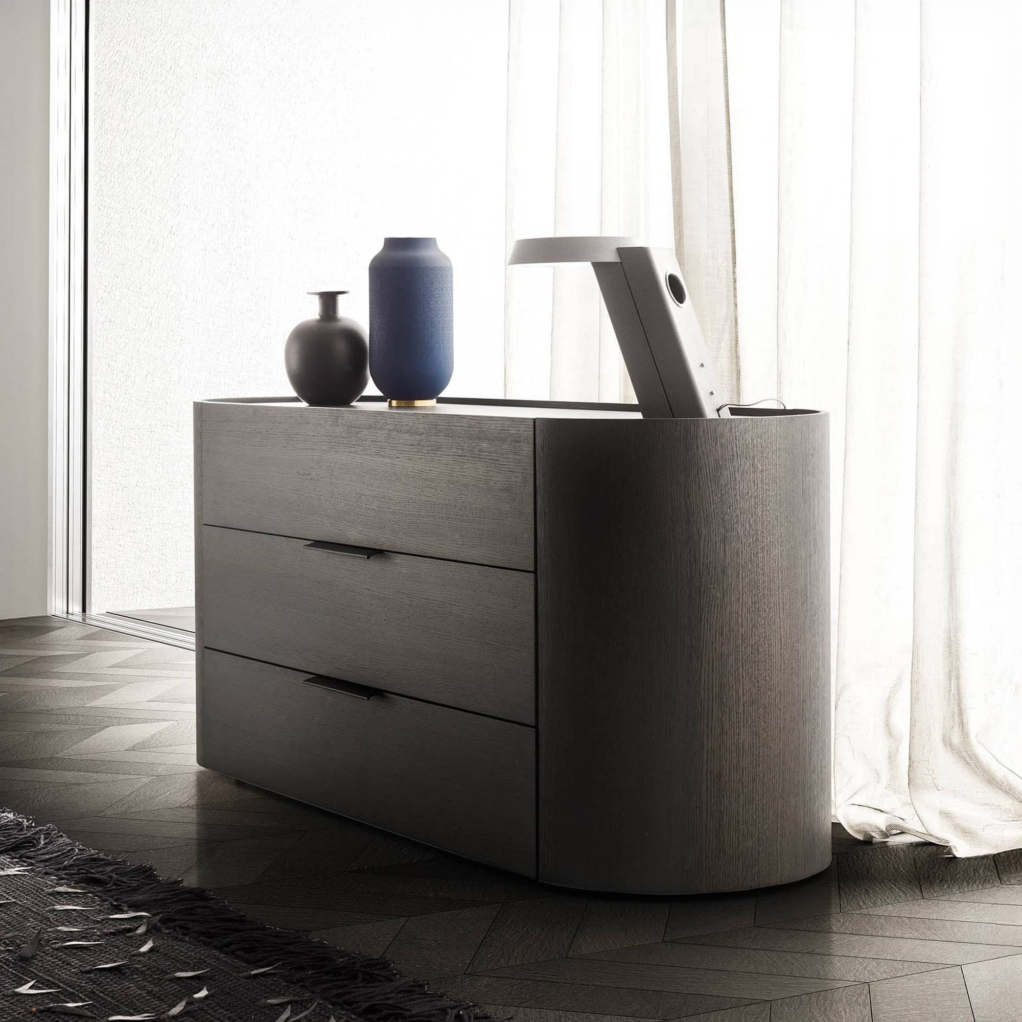 A dark wood dresser with rounded corners and three drawers, topped with two decorative vases and a pop-up light/screen. It sits on a dark herringbone wood floor next to sheer white curtains.