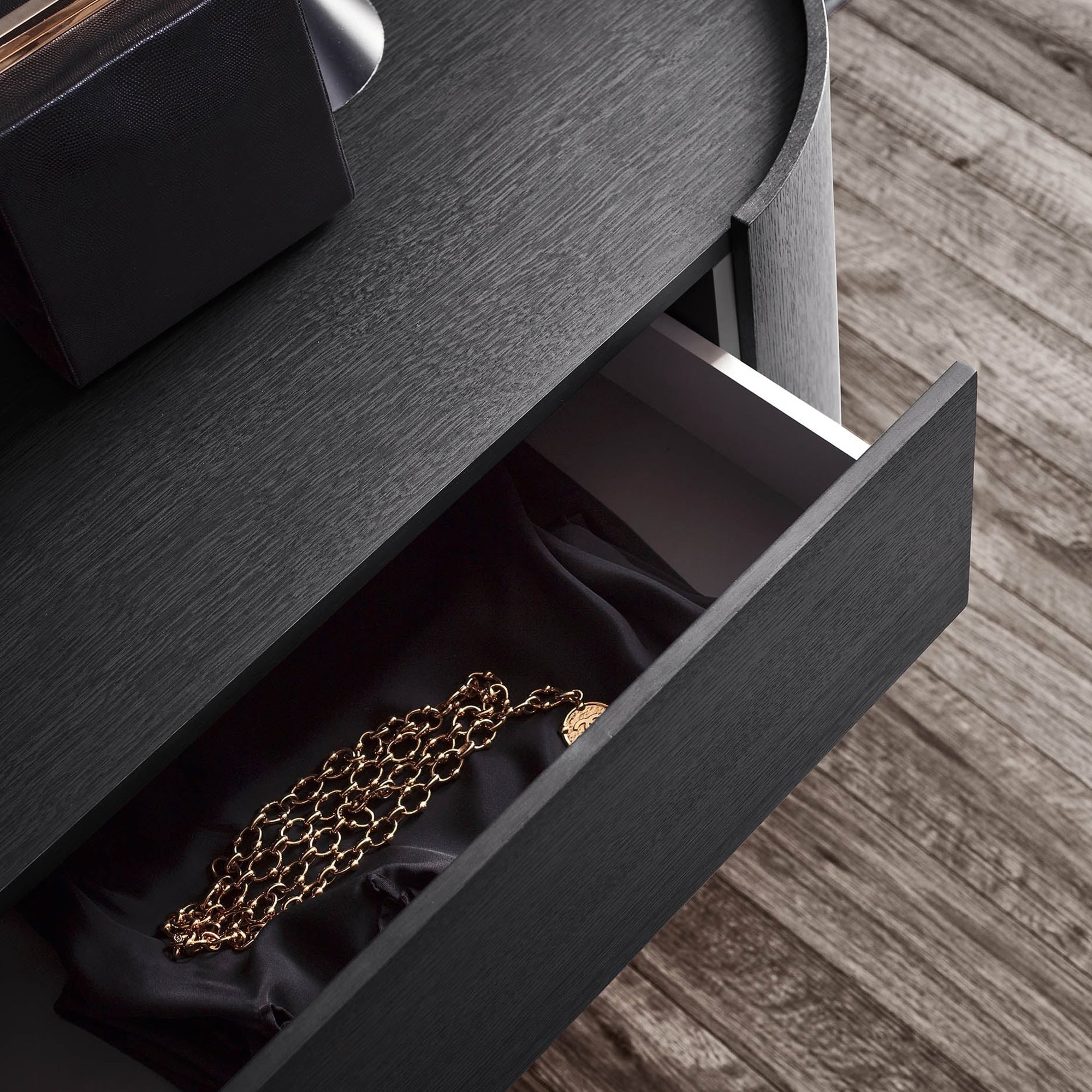 High-angle view of an open dark wood drawer, revealing a gold chain necklace with a coin pendant resting on black silk fabric. The dark wood table and a light wood floor are also visible.