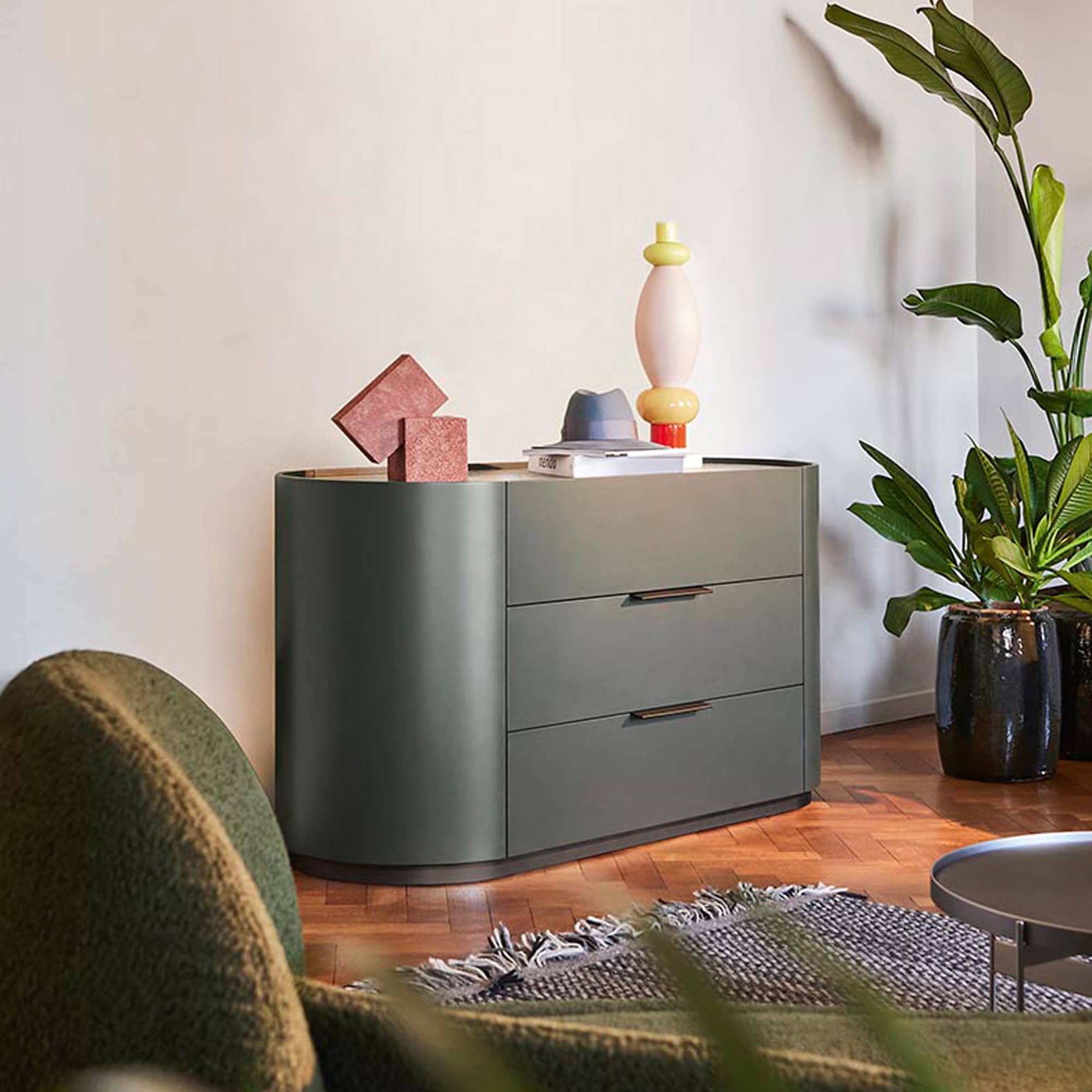 A modern olive-green dresser with rounded edges and three drawers, topped with decorative objects including a hat and abstract vase, next to a large potted plant in a contemporary room.