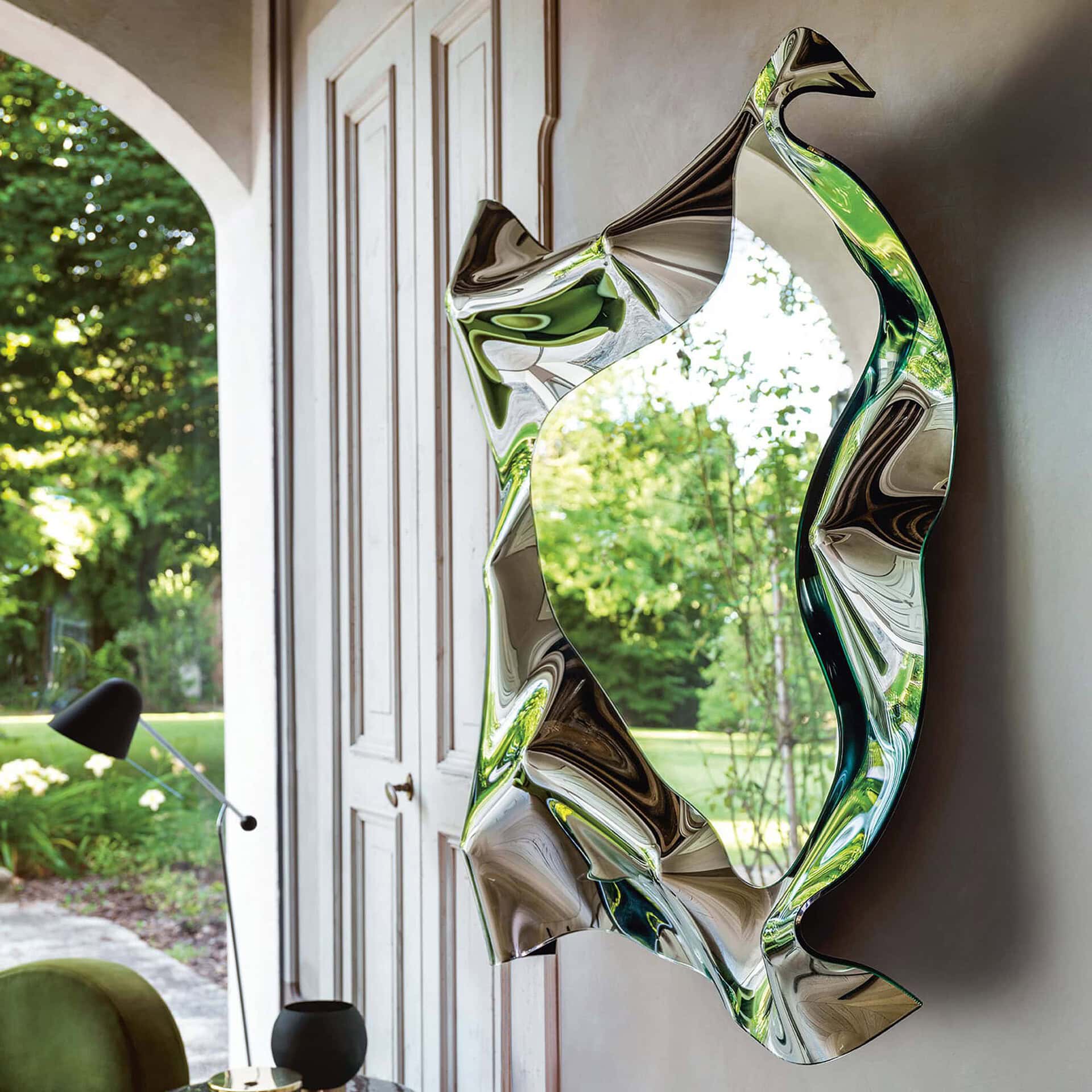Christine Mirror shown at an angle by a window, highlighting the fluid sculptural glass frame.