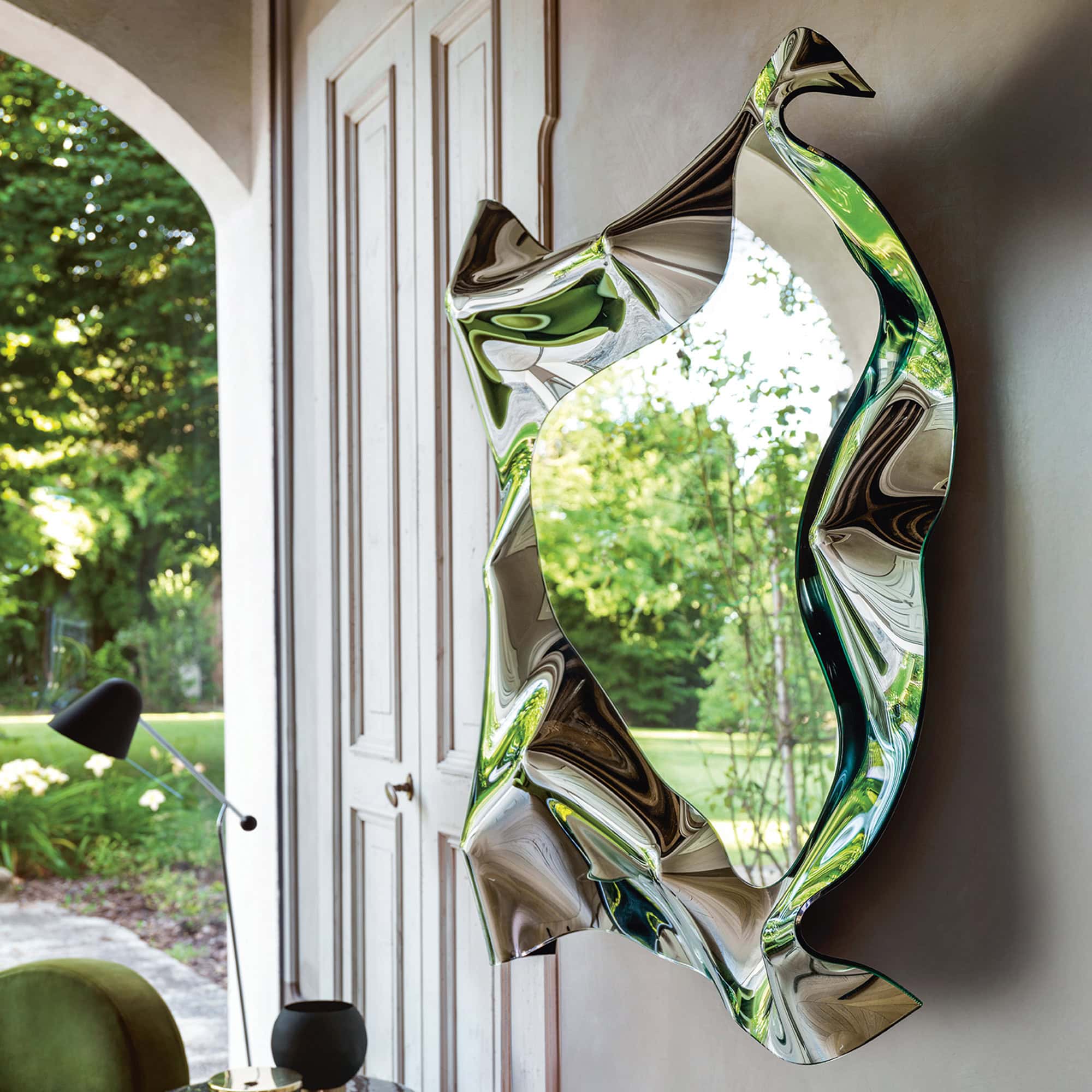 Christine Mirror shown at an angle by a window, highlighting the fluid sculptural glass frame.